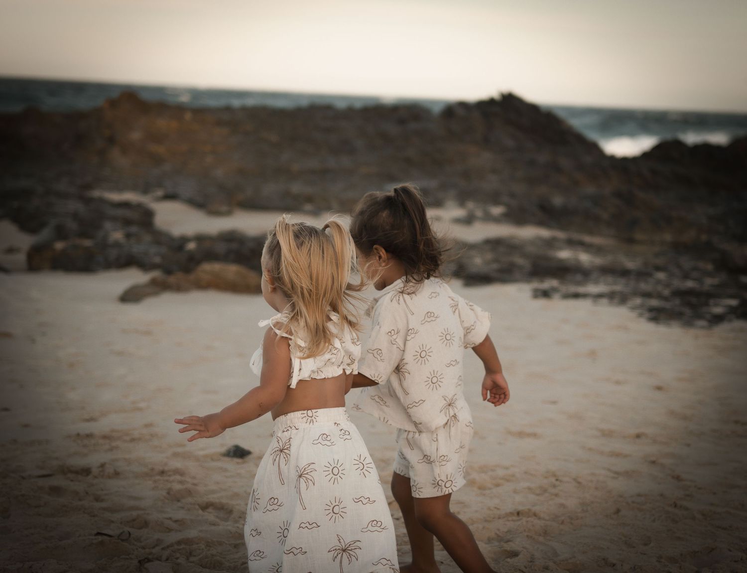 Two young girls in white outfits walking together on a sandy beach with rocky coastline at dusk.
