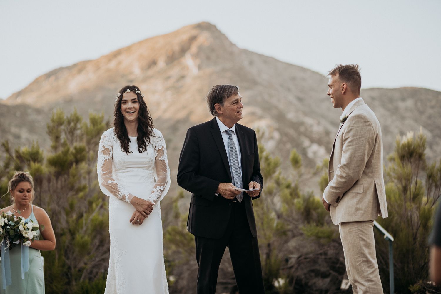 Outdoor wedding ceremony with bride in white lace gown, officiant, and groom in tan suit, set against a scenic mountain backdrop.