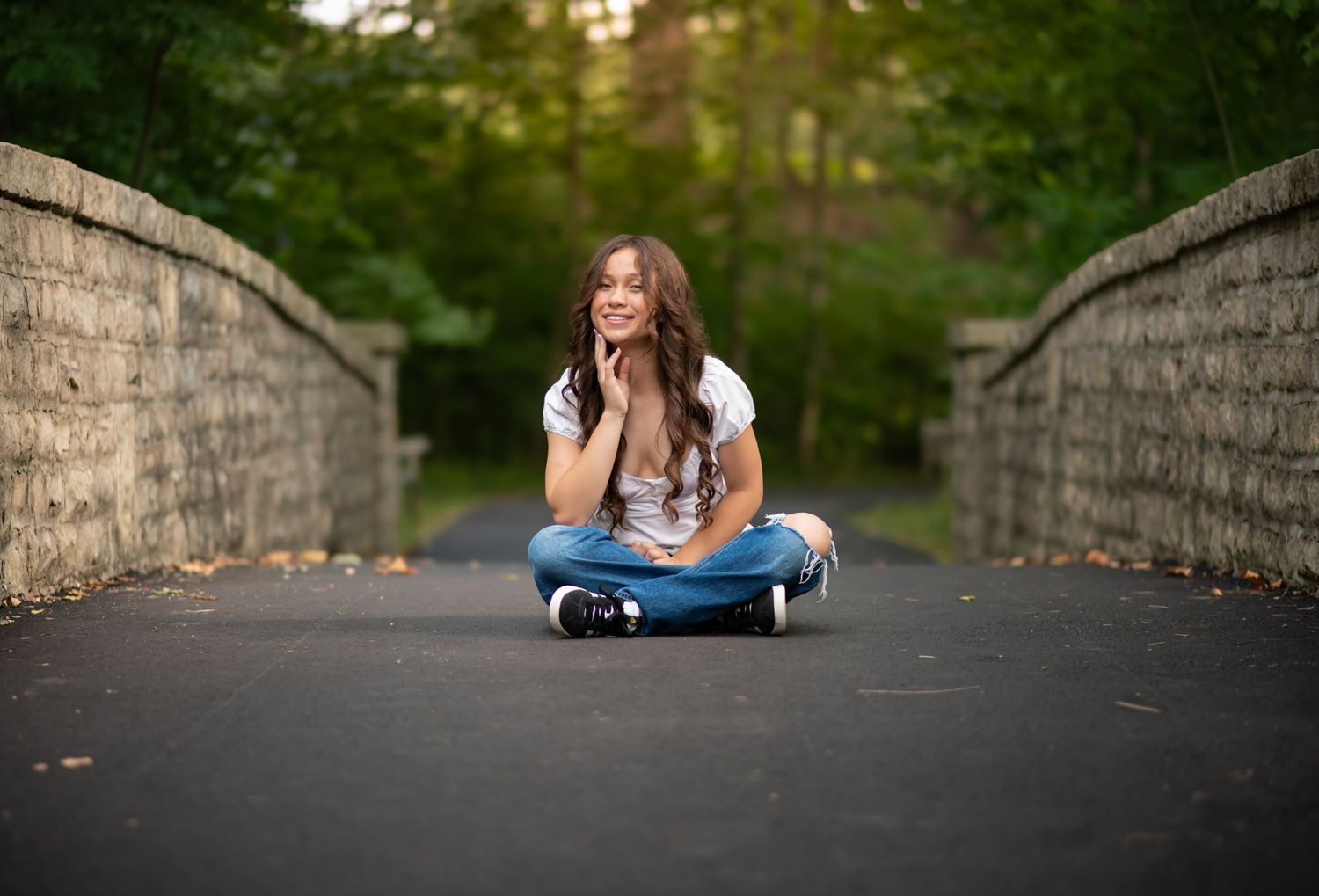 Young woman sitting cross-legged on a stone bridge surrounded by lush green trees, smiling.