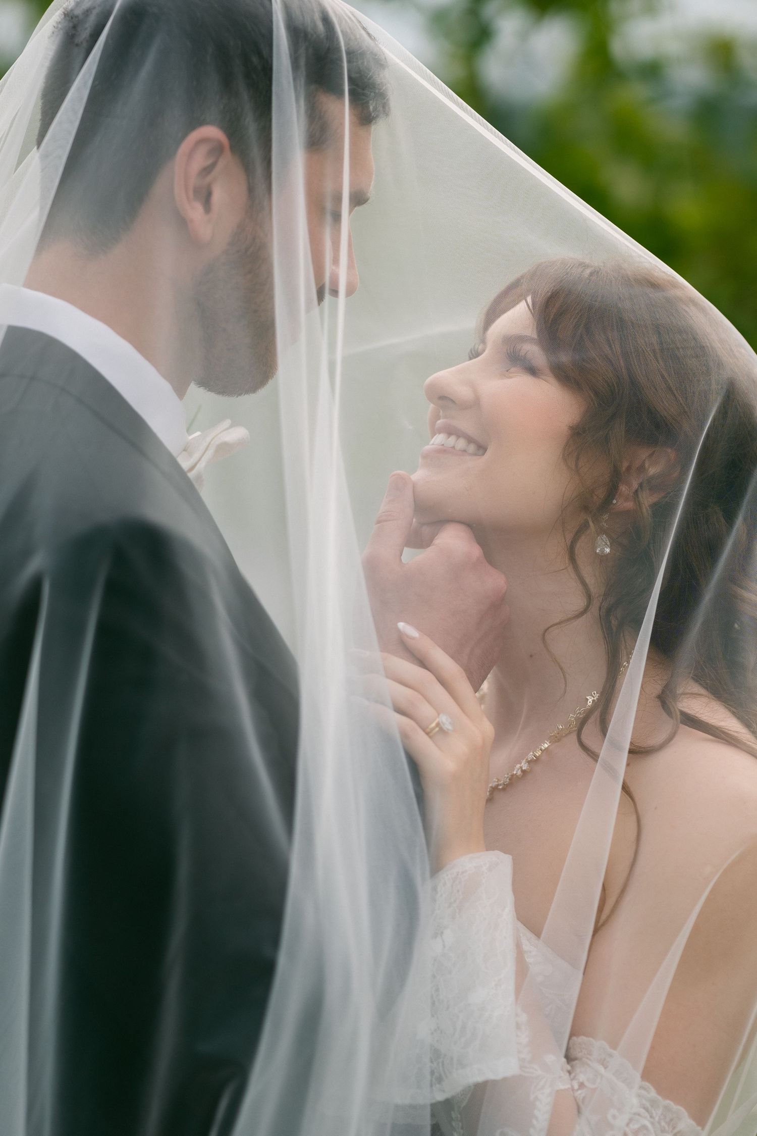 Bride and groom share a romantic moment under a wedding veil, with the smiling bride gazing up at her groom.