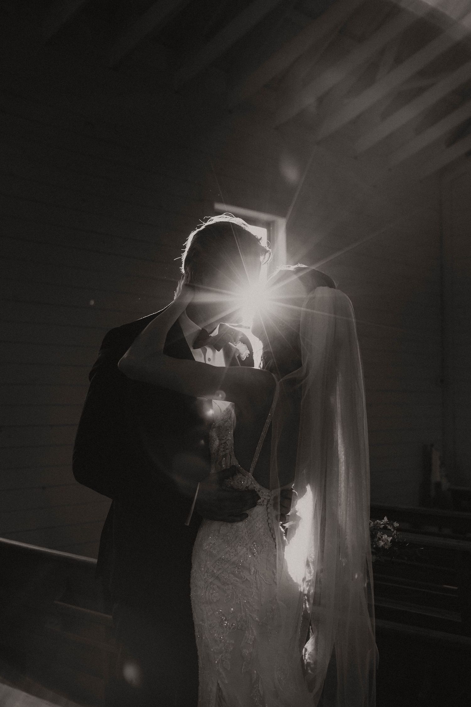 Black and white wedding photo of bride and groom embracing in church with dramatic sunburst light through window.