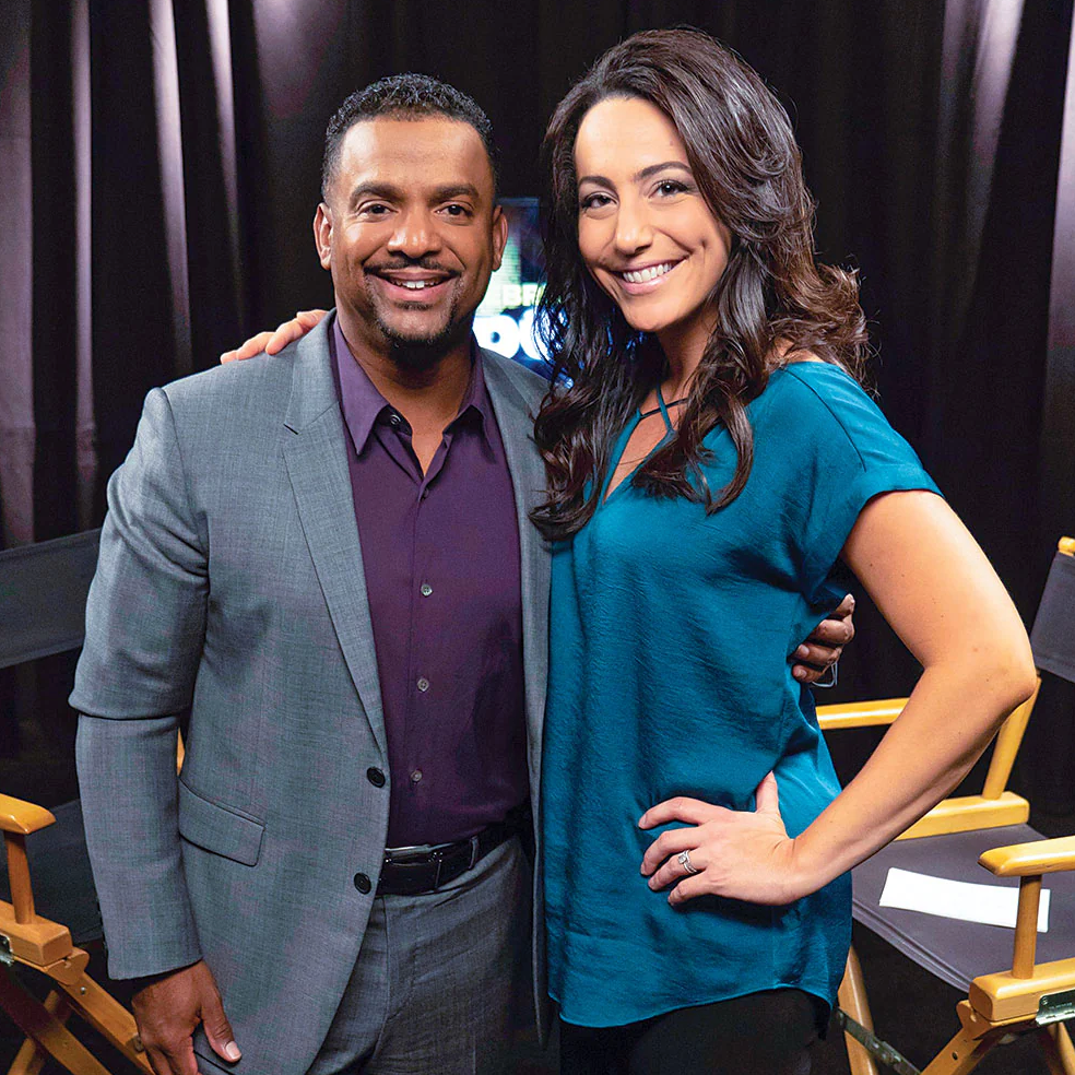 Two people in black outfits smiling and posing together in front of director chairs on a white background.