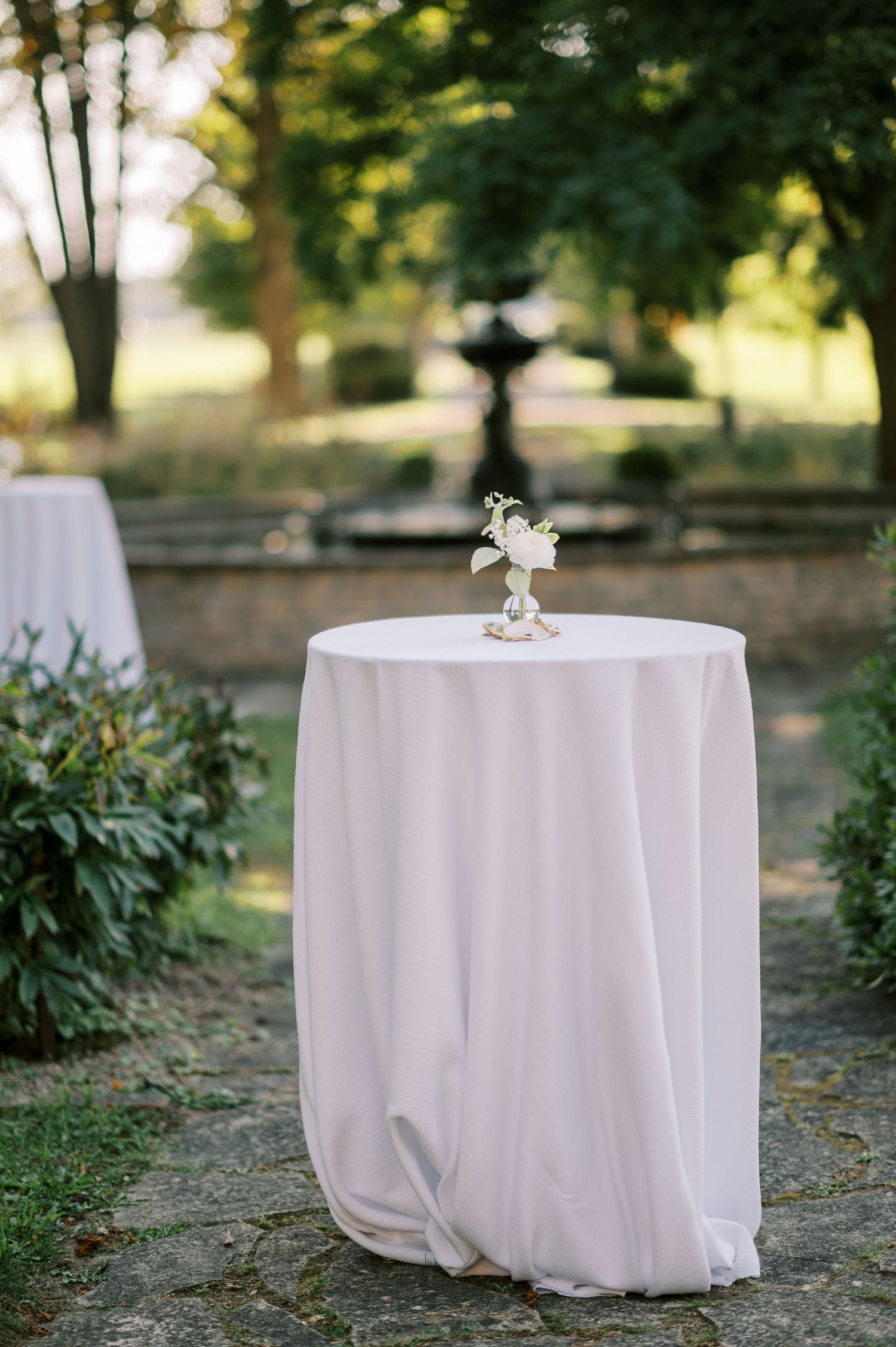 Elegant outdoor cocktail hour setup with a white draped high-top table topped with a small floral arrangement in a garden setting.