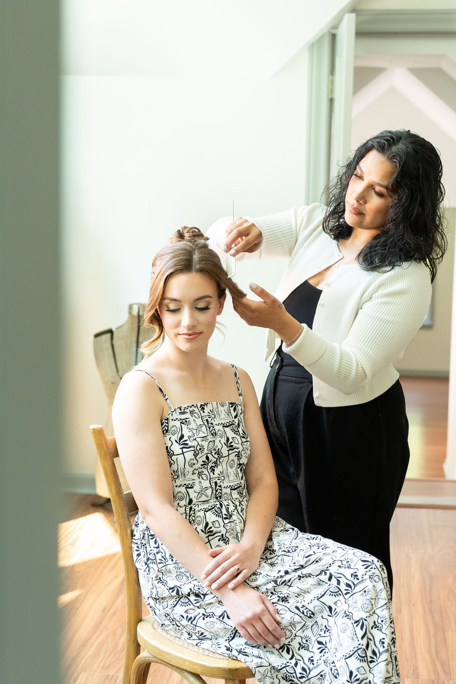 A hairstylist styling a young woman's updo in a bright salon, both wearing elegant black and white outfits.