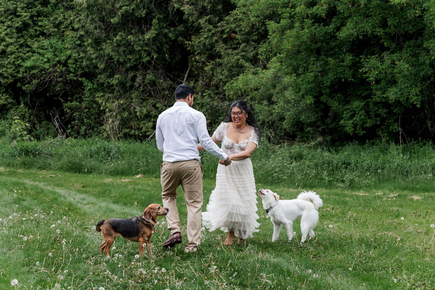 Candid engagement photo of couple hold hands while spinning in a field with their two dogs in the guelph Arboretum.