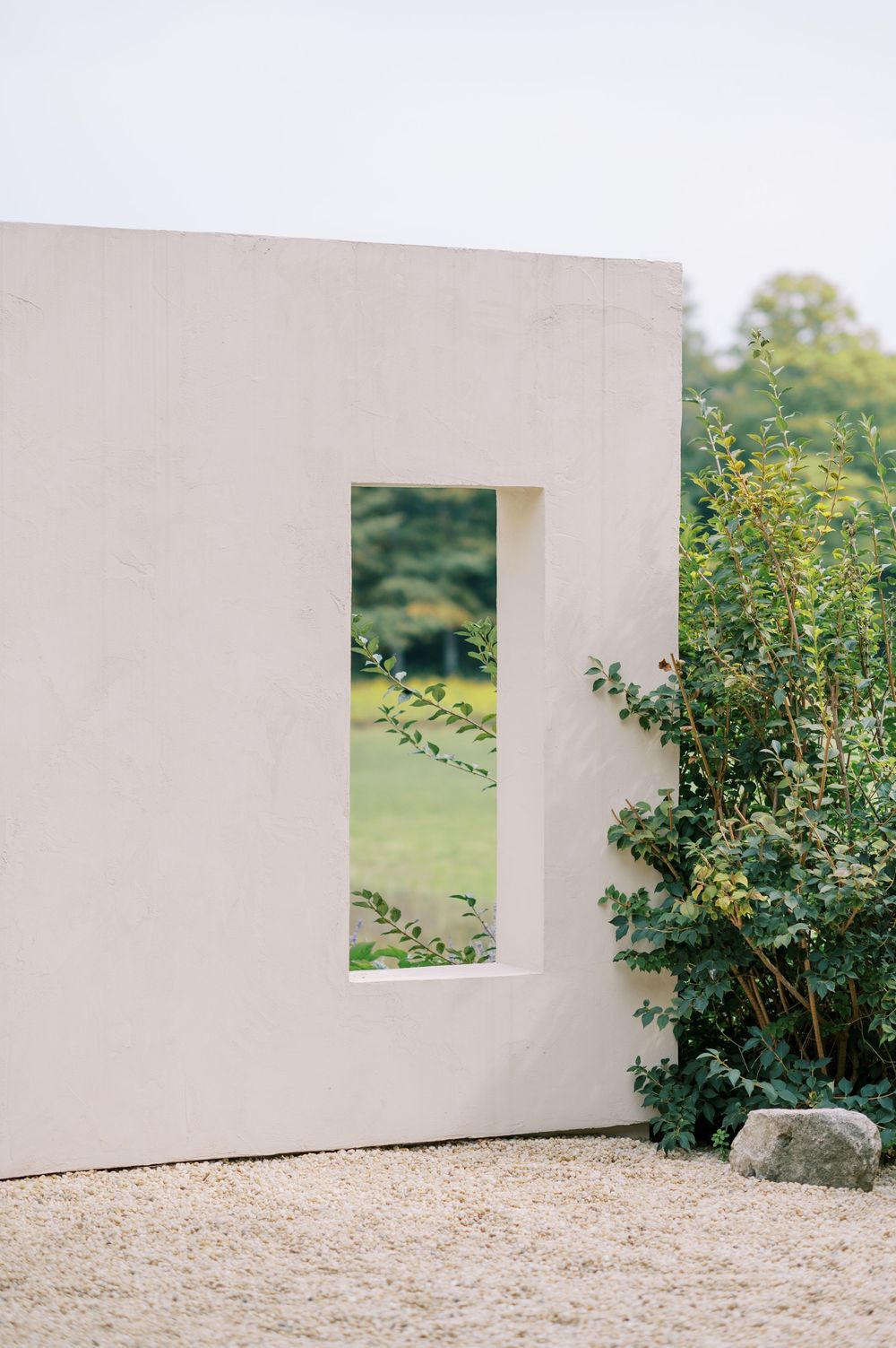 White minimalist garden wall with rectangular window opening revealing lush green landscape beyond, flanked by climbing roses.