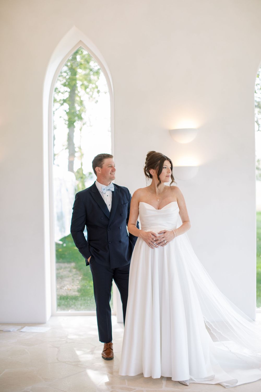 Bride in strapless white ball gown and groom in navy suit stand together in a bright white chapel with arched windows.