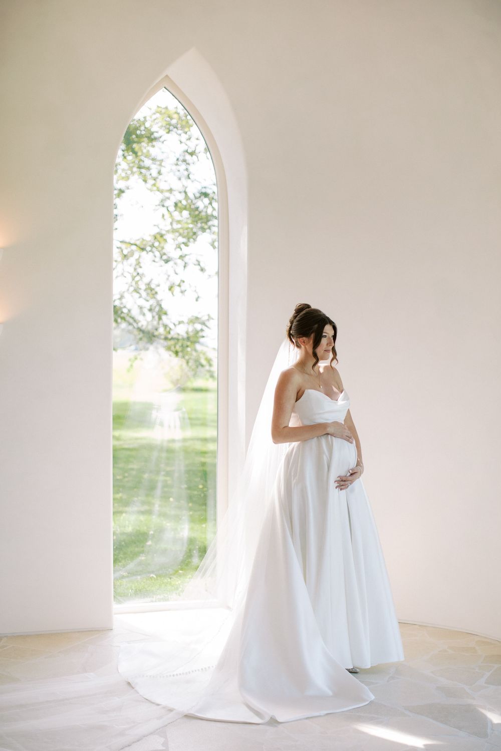 Pregnant bride in white strapless gown and veil stands by arched chapel window in soft natural light.