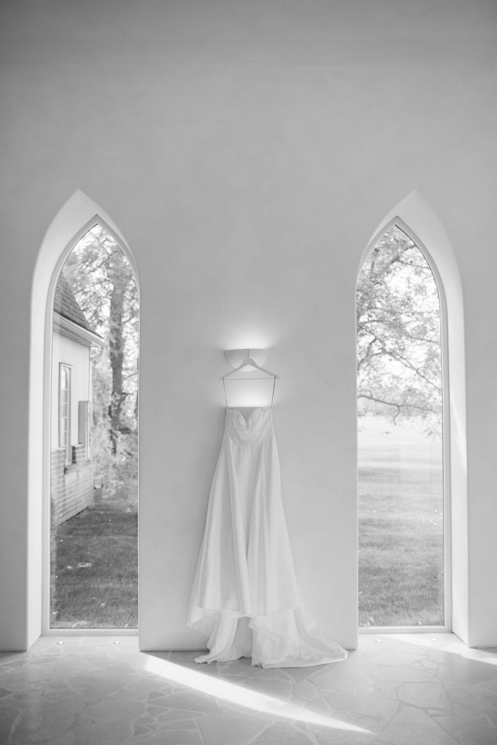 Black and white photo of a wedding dress hanging between two Gothic arch windows in a white chapel.