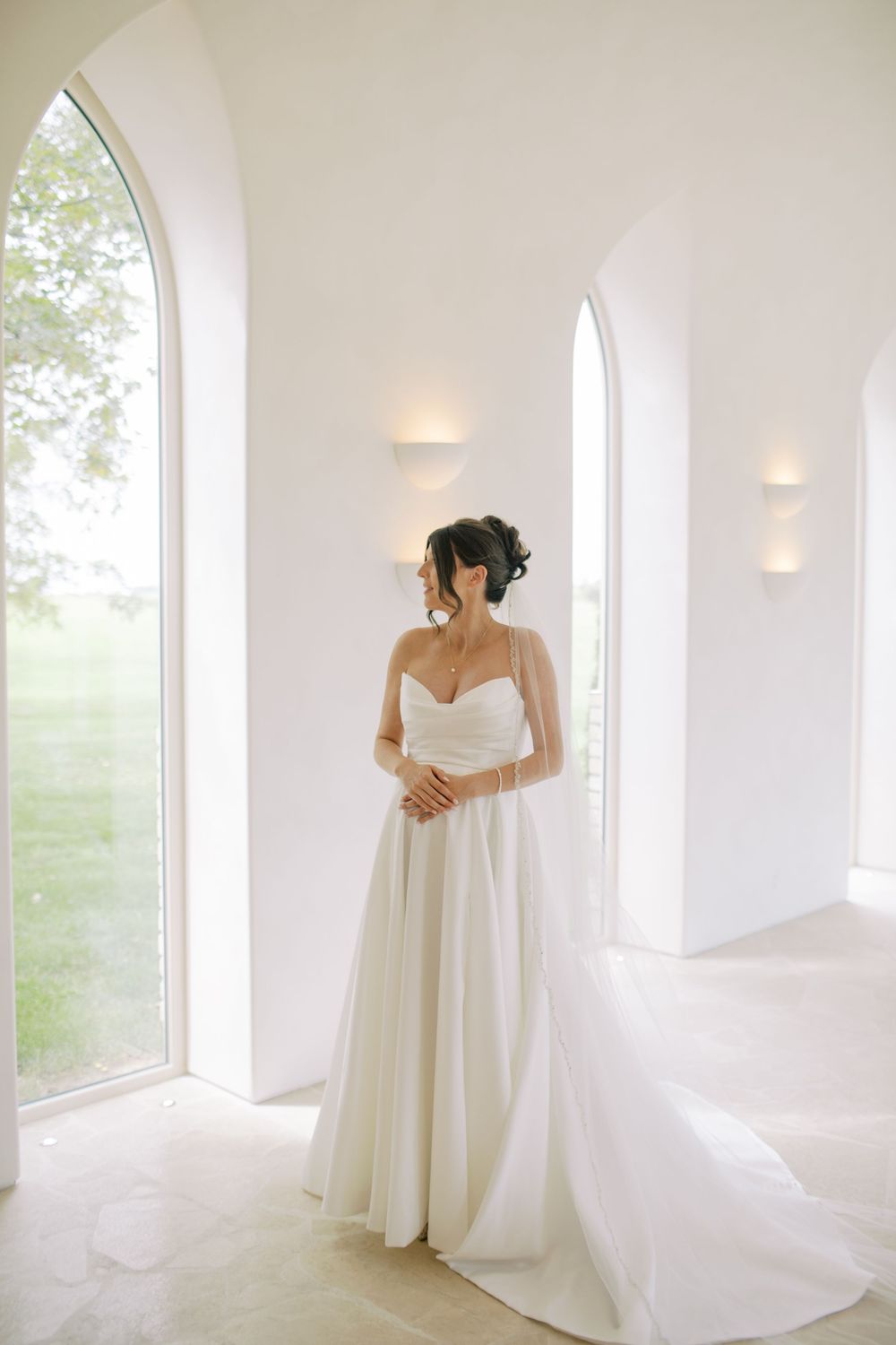 Elegant bride in a white strapless ball gown stands in a bright white chapel with arched windows and soft wall sconces.