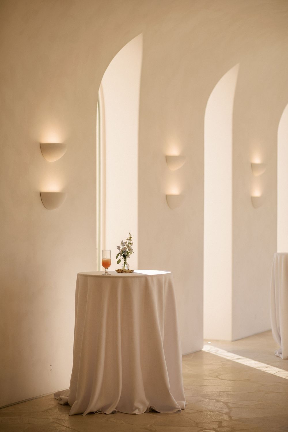 Elegant white cocktail table with floral arrangement and wine glass in arched hallway with warm wall sconces.