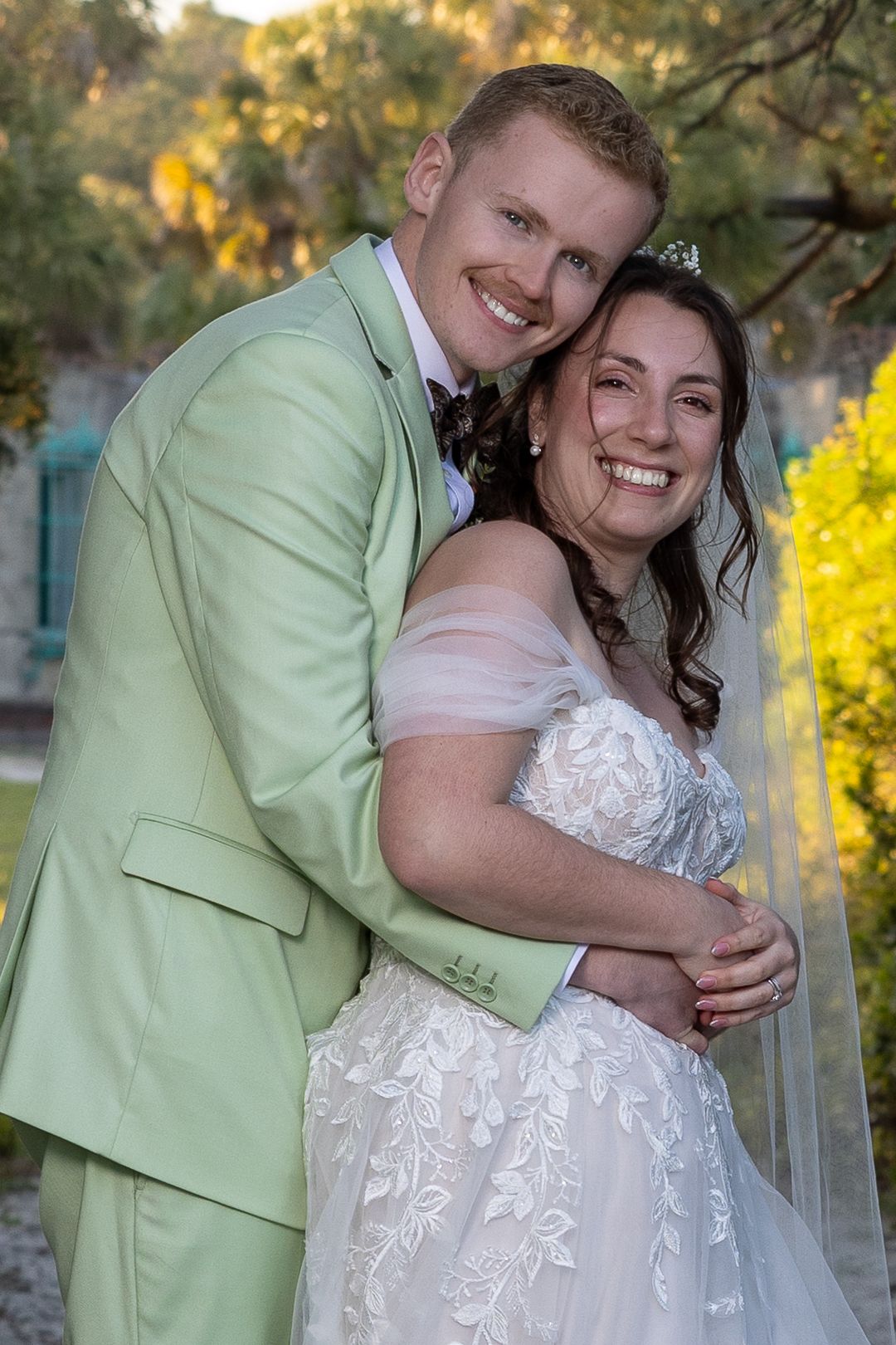 Smiling bride in lace gown and groom in mint green suit embrace outdoors at their wedding ceremony.