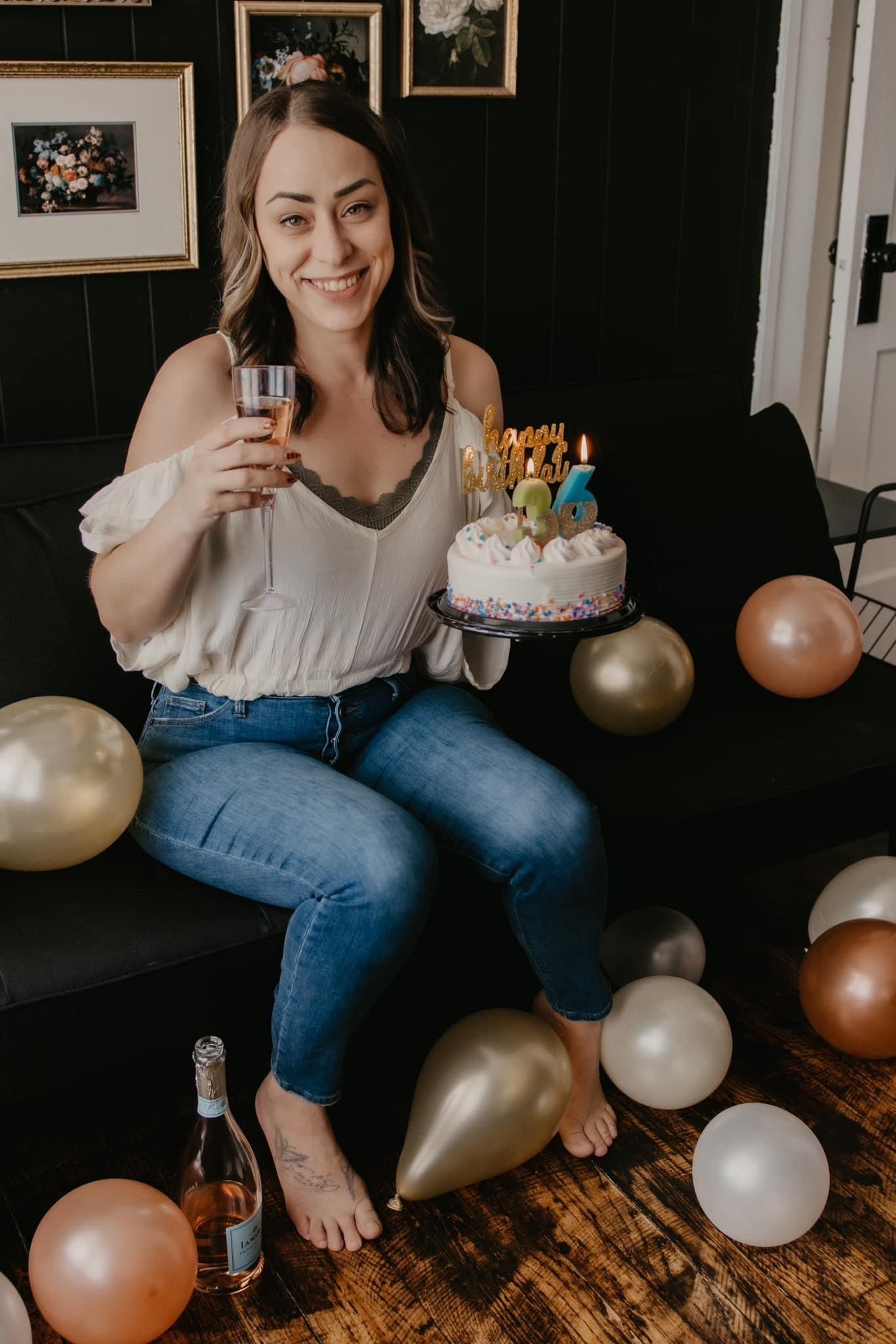 Woman celebrates birthday sitting on sofa with cake, champagne, and gold and rose gold balloons.