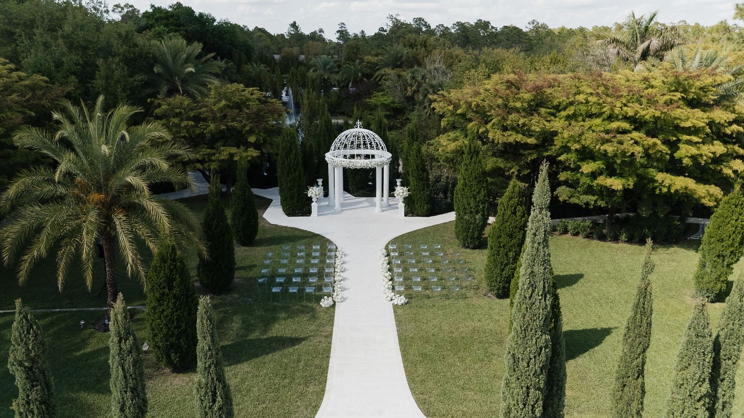 Aerial view of elegant white gazebo wedding venue with manicured cypress trees and palm trees lining a white aisle.
