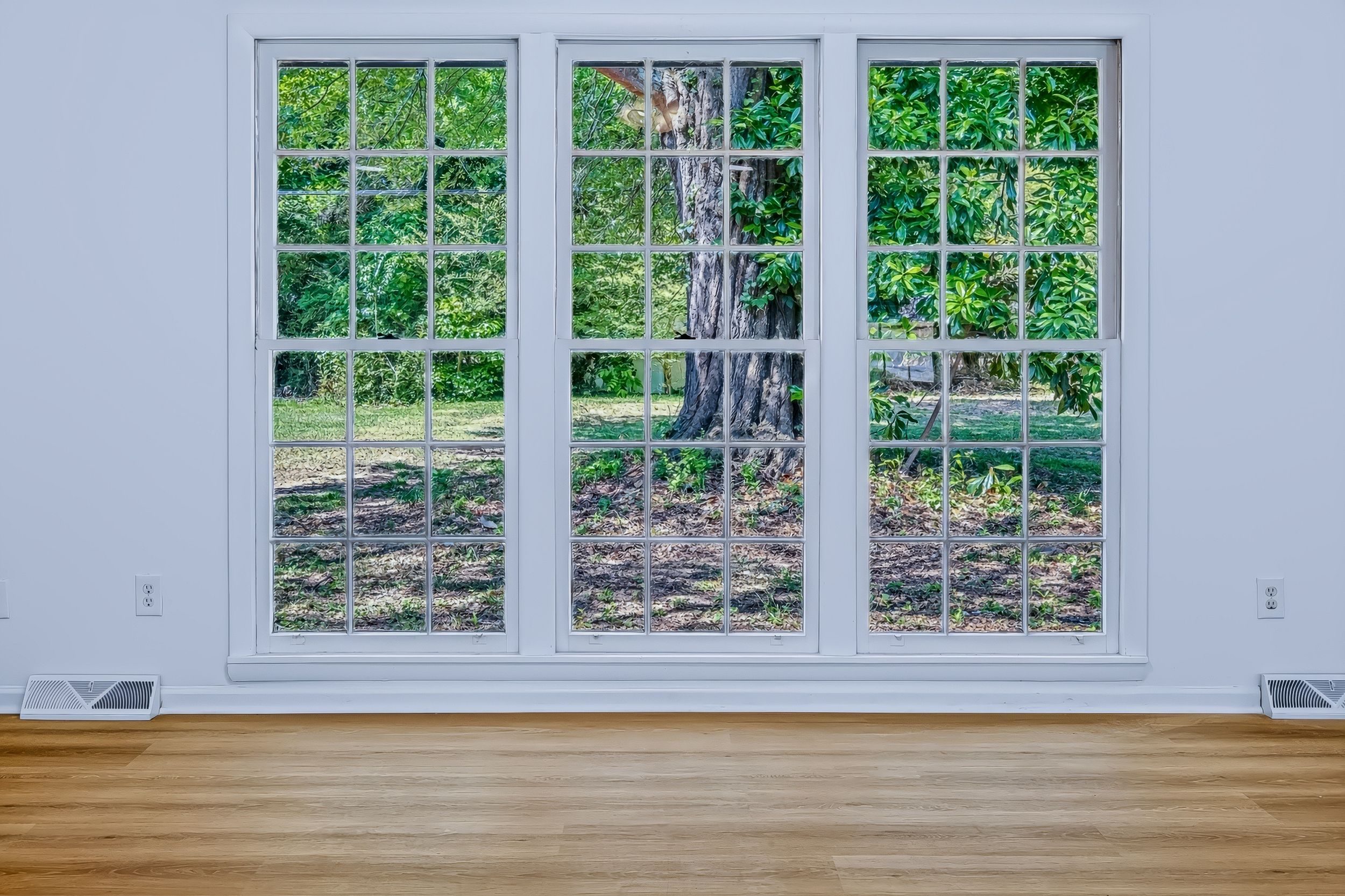 Empty room with hardwood floors and three large white-framed windows overlooking lush green trees and garden.