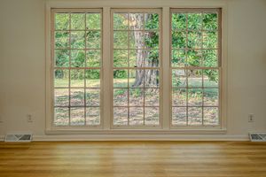 Large triple-pane windows with white trim overlooking lush green trees on hardwood floors with floor vents.