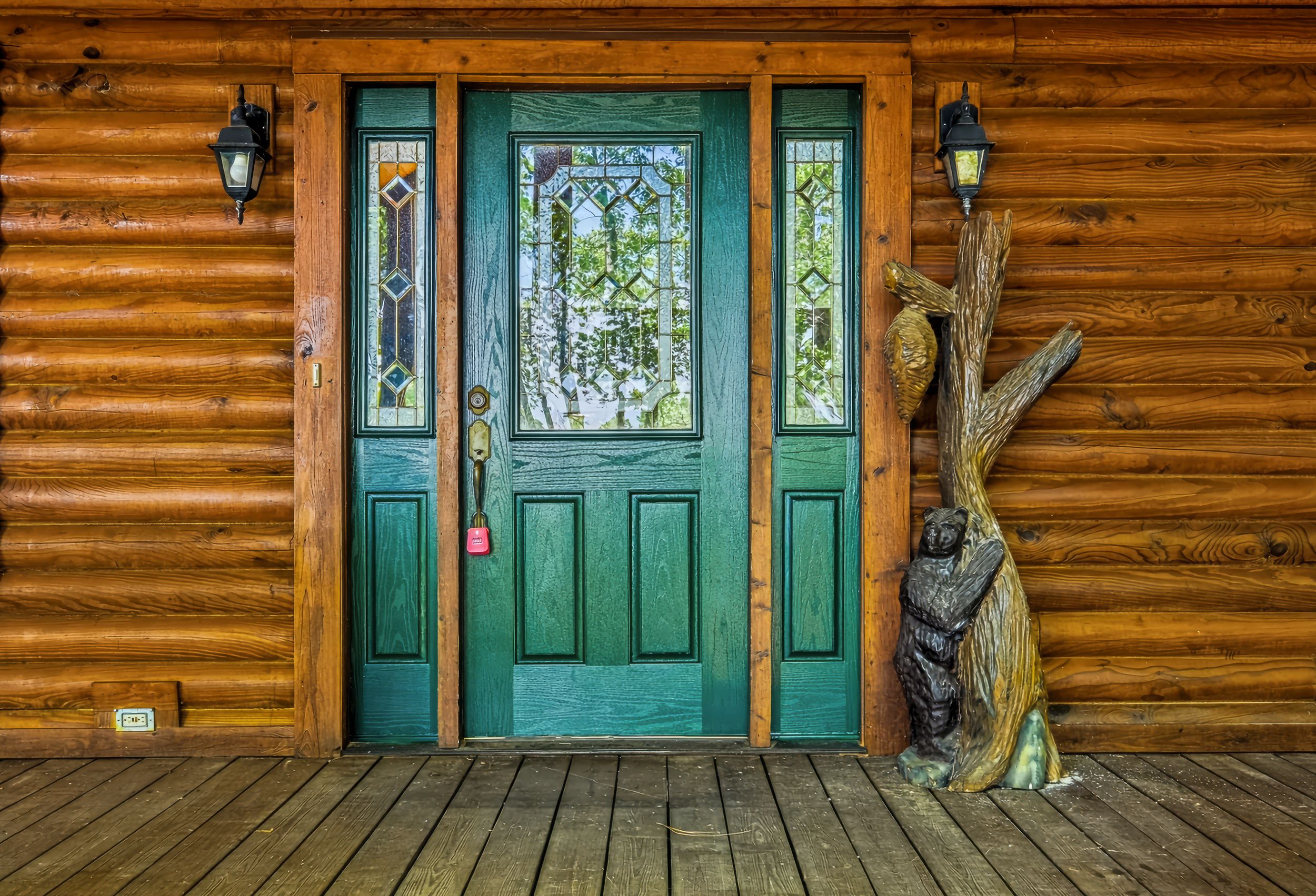 Rustic log cabin entrance with teal stained-glass door, wood carved bear sculpture, and lantern wall sconces on a wooden porch.