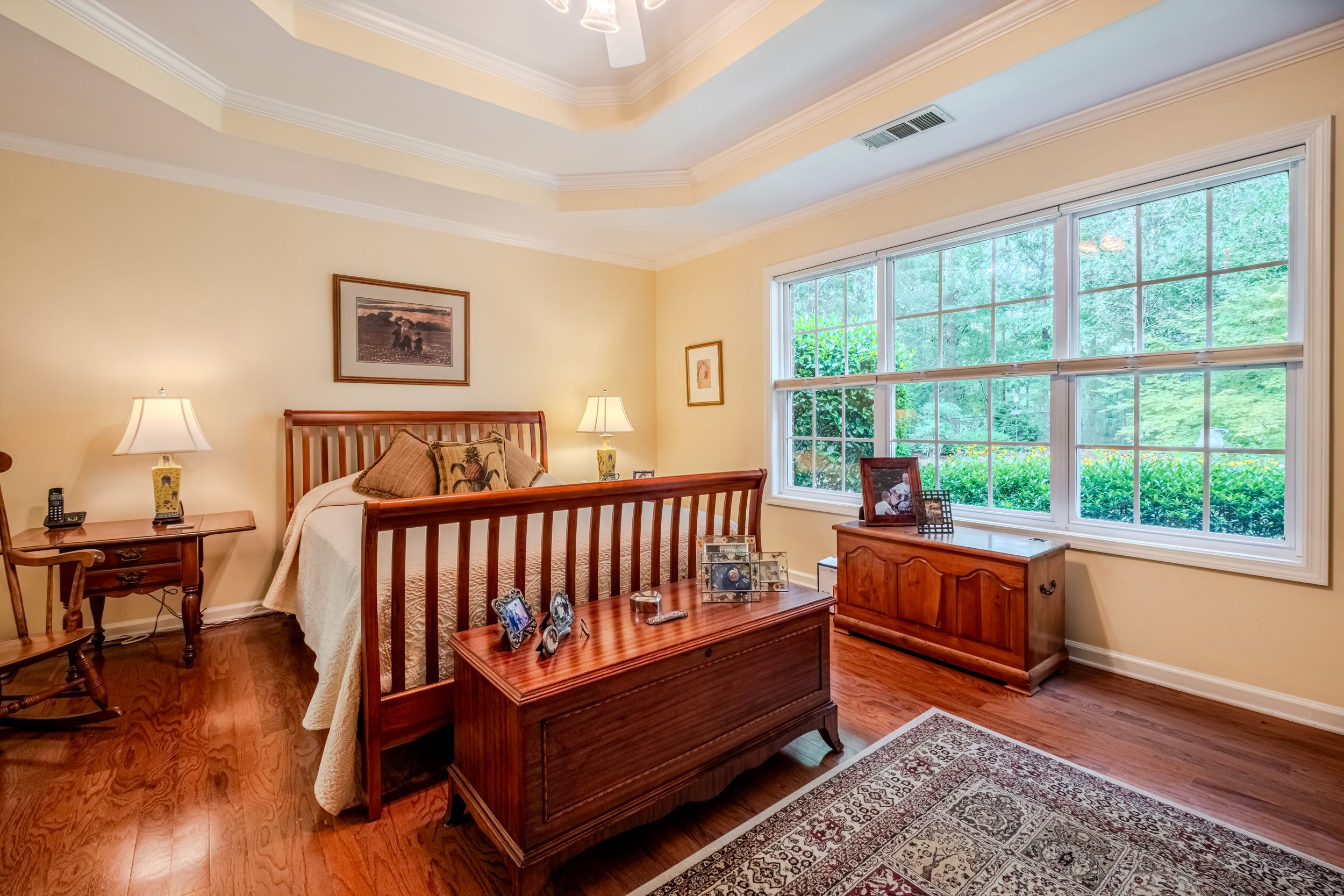 Elegant master bedroom with hardwood floors, tray ceiling, wooden sleigh bed, antique chest, and large windows overlooking lush greenery.
