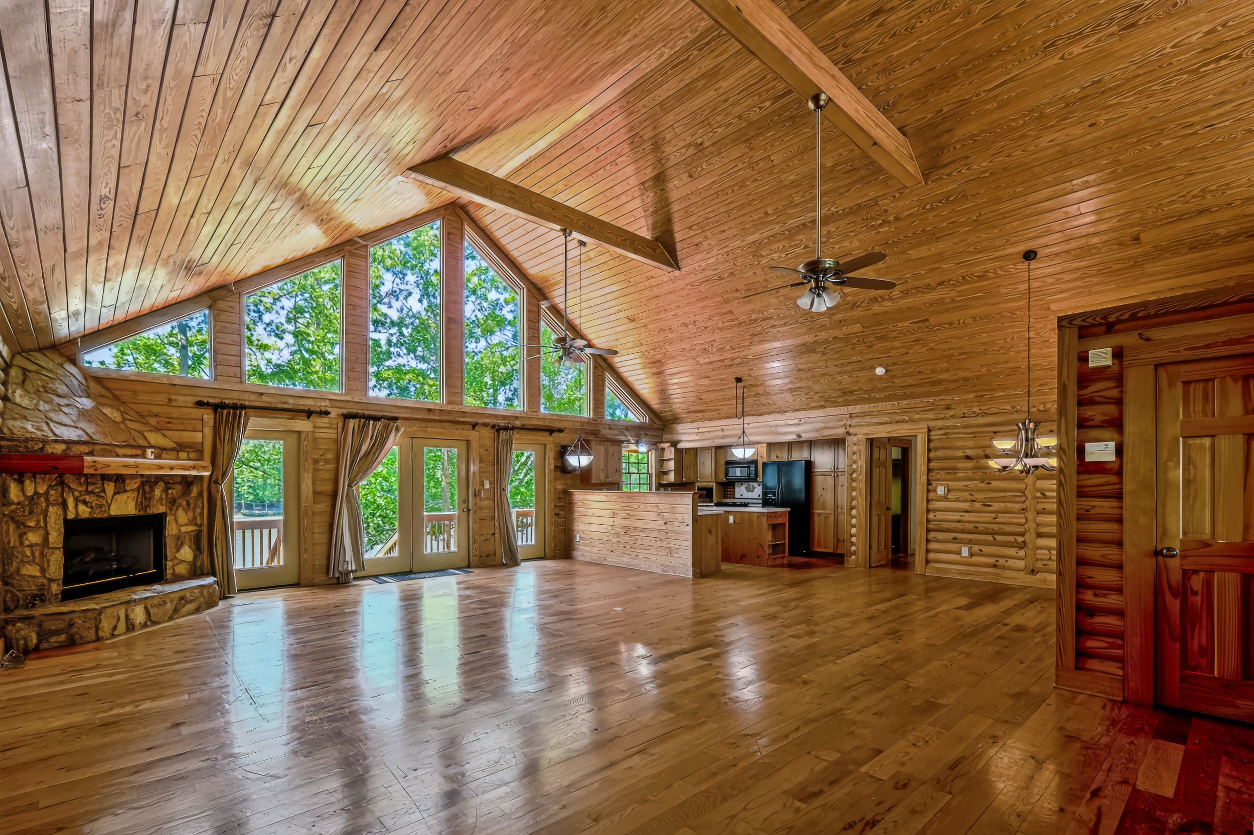 Spacious log cabin great room with vaulted wood ceiling, hardwood floors, stone fireplace, and large windows overlooking trees.