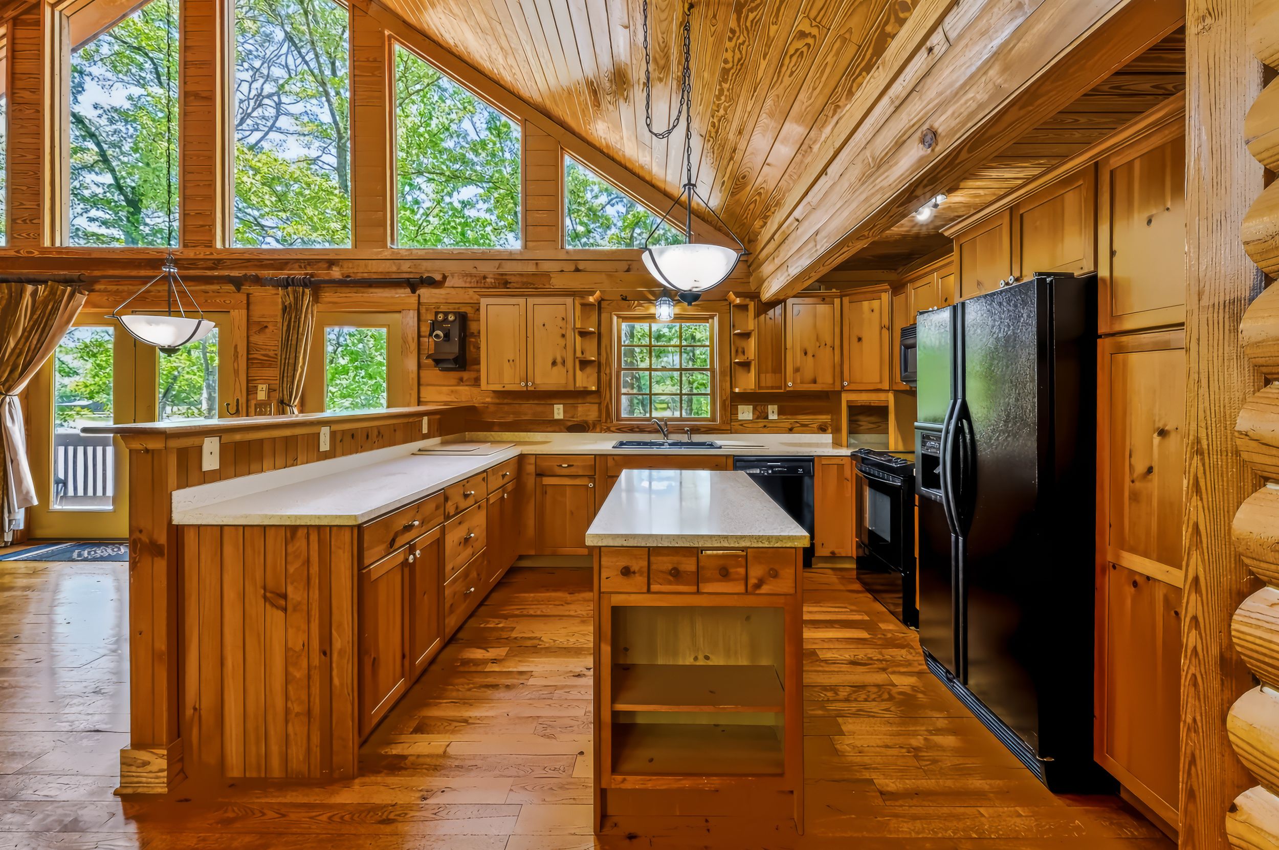 Rustic log cabin kitchen with wood cabinetry, center island, hardwood floors, and black appliances surrounded by large windows.