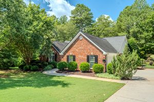 Brick ranch home with manicured lawn, lush green trees, dark shutters, and a concrete driveway in a wooded suburban setting.