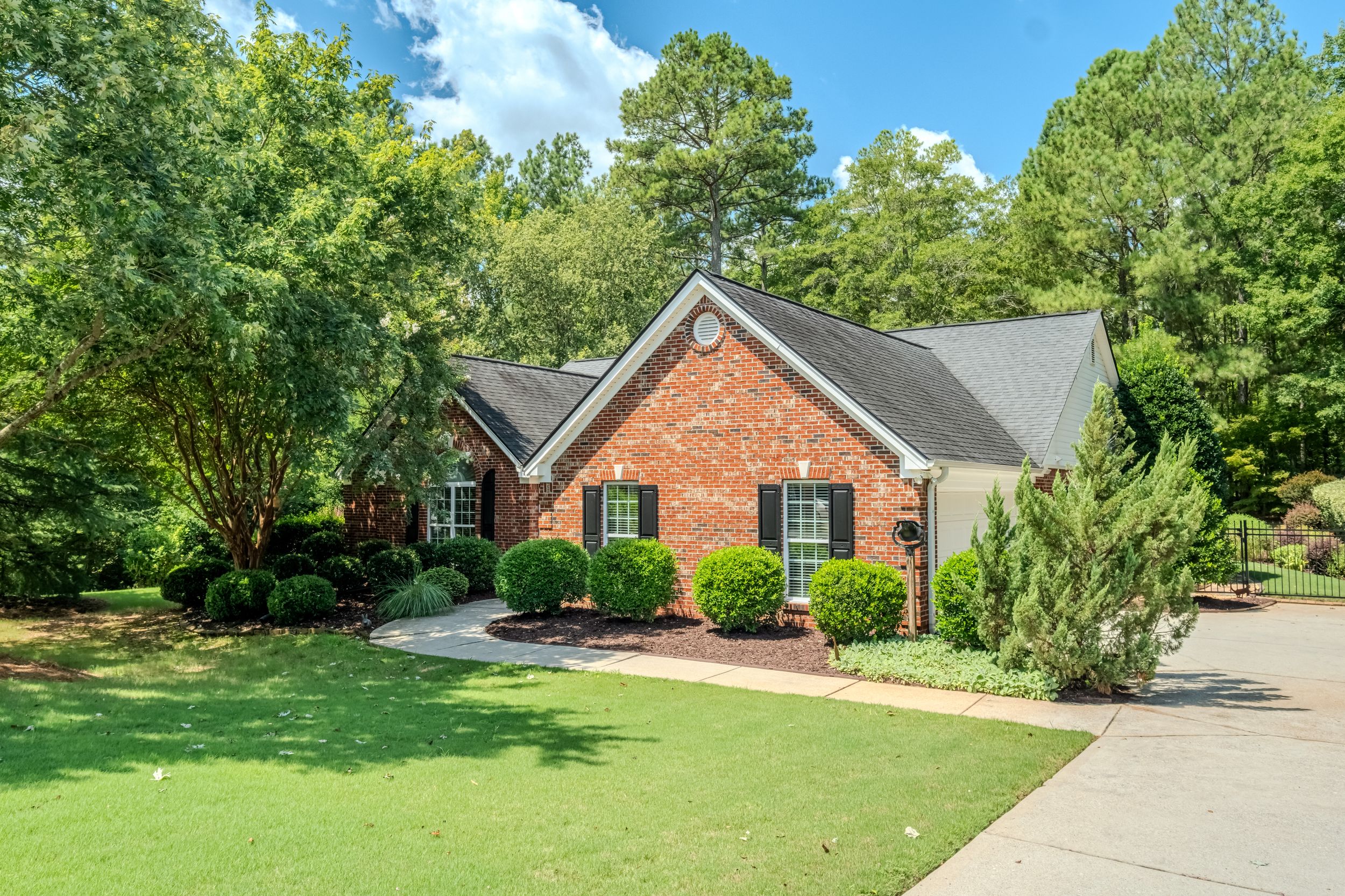 Brick ranch home with manicured lawn, lush green trees, dark shutters, and a concrete driveway in a wooded suburban setting.