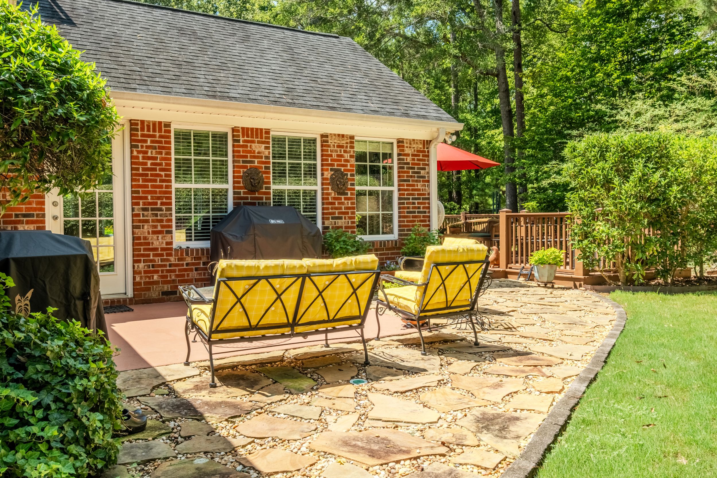 Brick cottage with stone patio featuring yellow cushioned chairs, grill, and red umbrella surrounded by lush trees.
