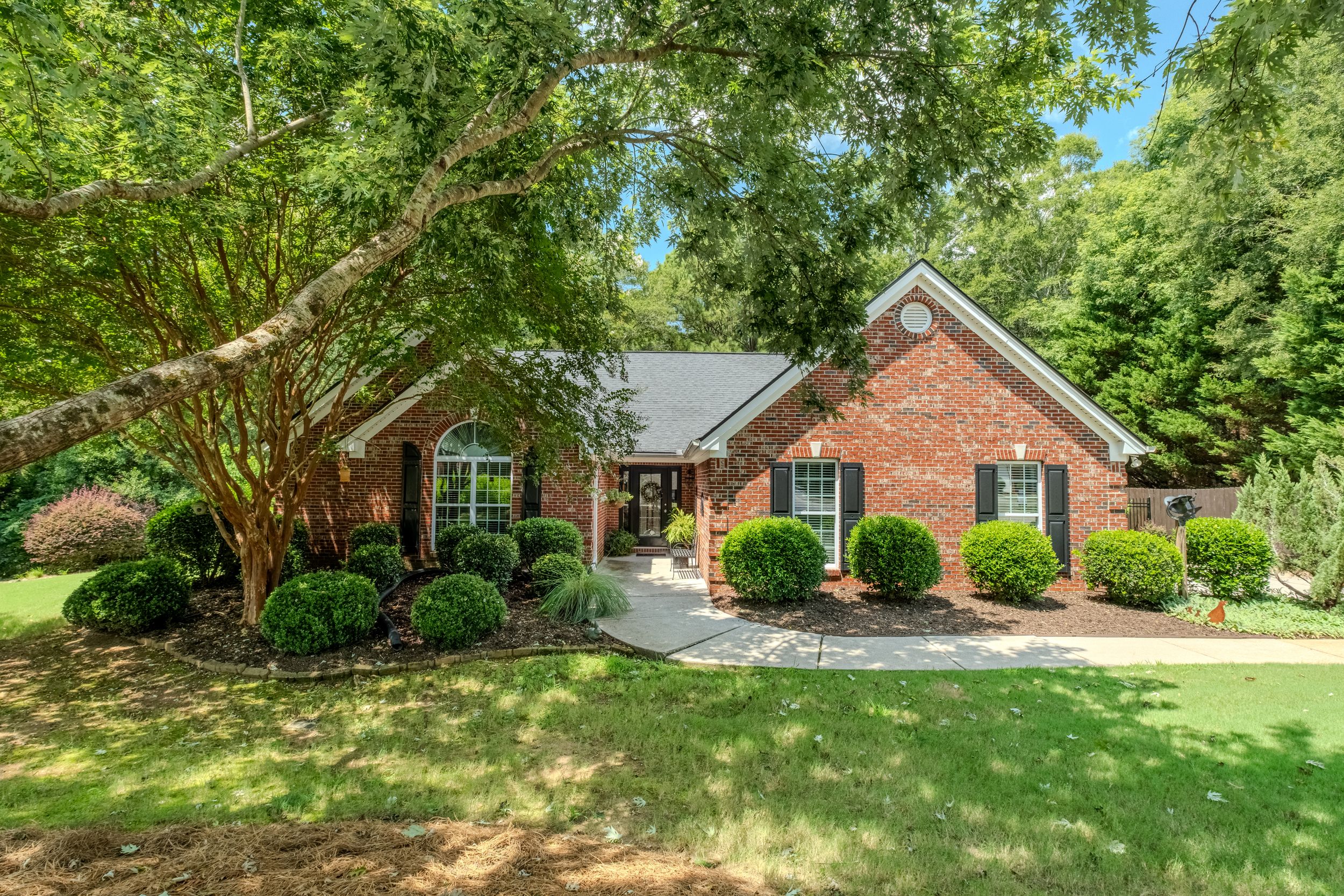 Brick ranch home with lush green lawn, mature trees, manicured shrubs, black shutters, and charming curb appeal.