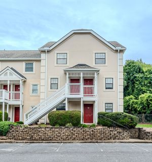 Three-story beige townhouse with red doors, white staircase, and balcony, surrounded by landscaping and parking area.