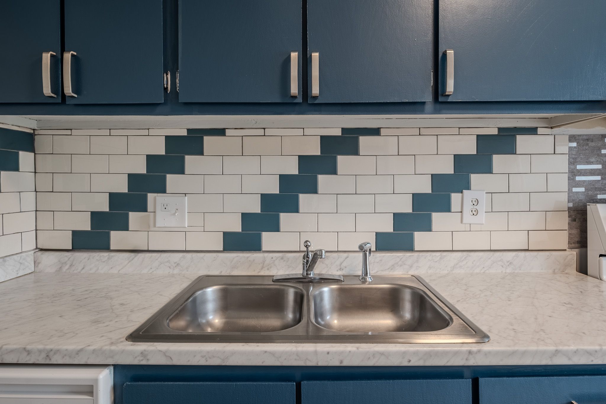 Modern kitchen sink with double basin, blue cabinets, and white subway tile backsplash with blue diagonal accent pattern.