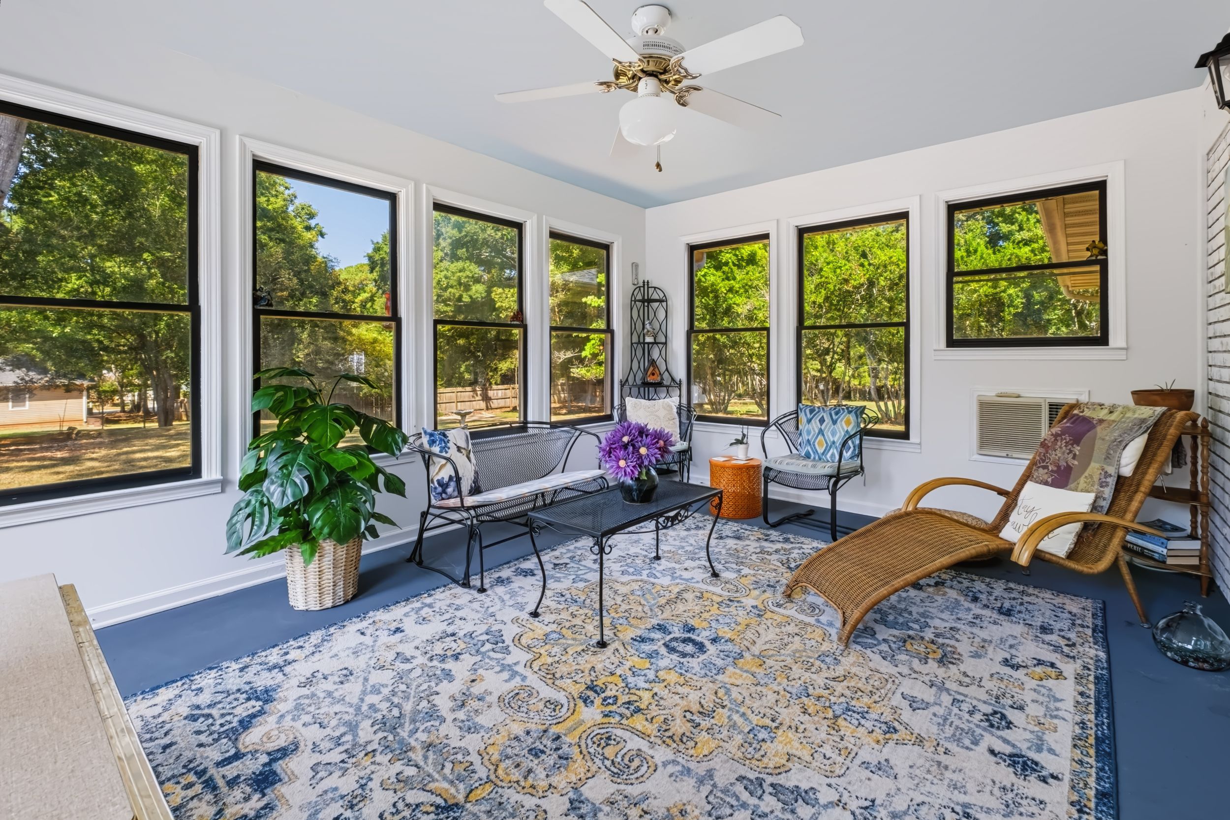 Bright sunroom with large windows, blue floral area rug, wicker chairs, and lush garden views.