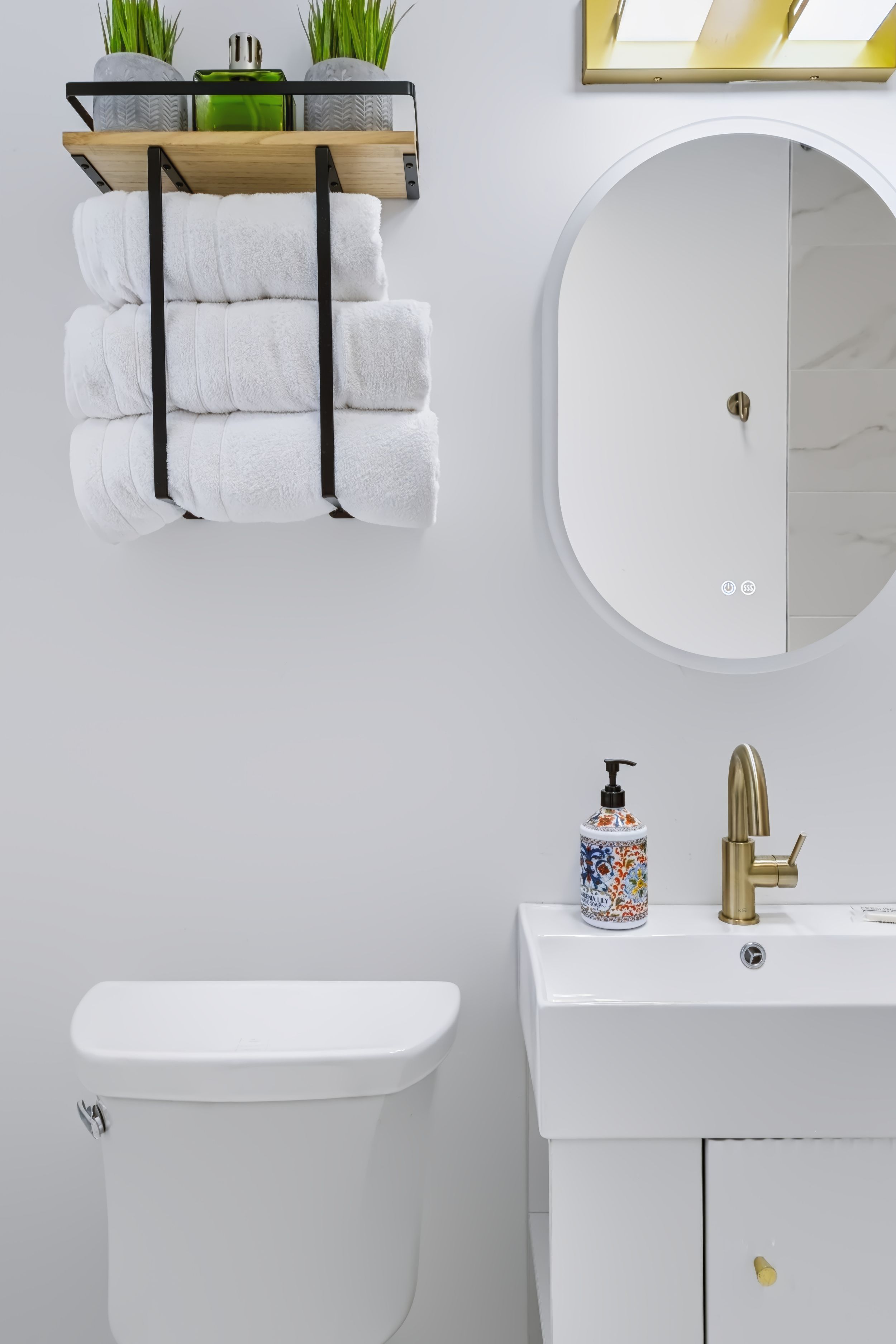 Modern white bathroom with oval mirror, gold faucet, towel rack, and decorative soap dispenser on white vanity.