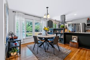 Bright dining area with round table, gray chairs, hardwood floors, area rug, and large windows overlooking lush greenery.