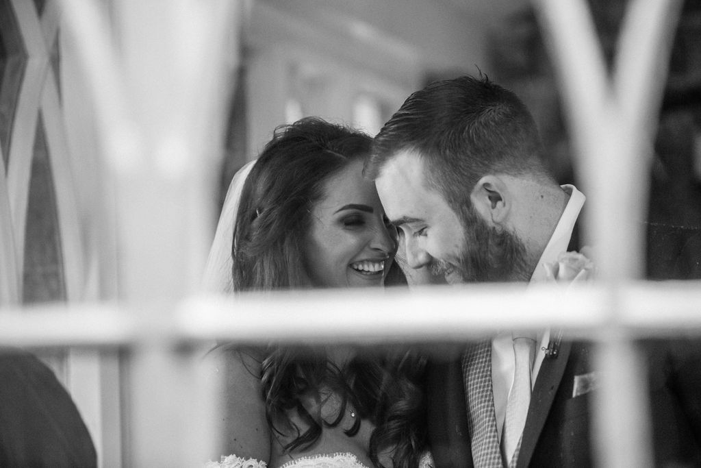 Black and white photo of a smiling bride and bearded groom sharing an intimate moment on their wedding day.