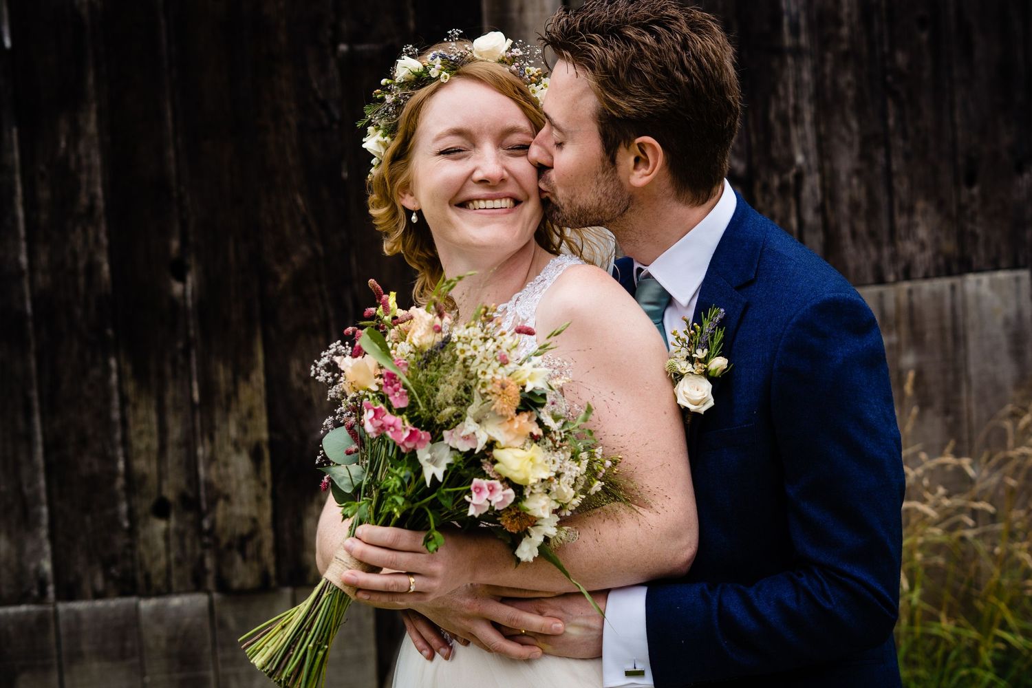 Bride with floral crown holds bouquet as groom kisses her cheek near rustic barn backdrop.