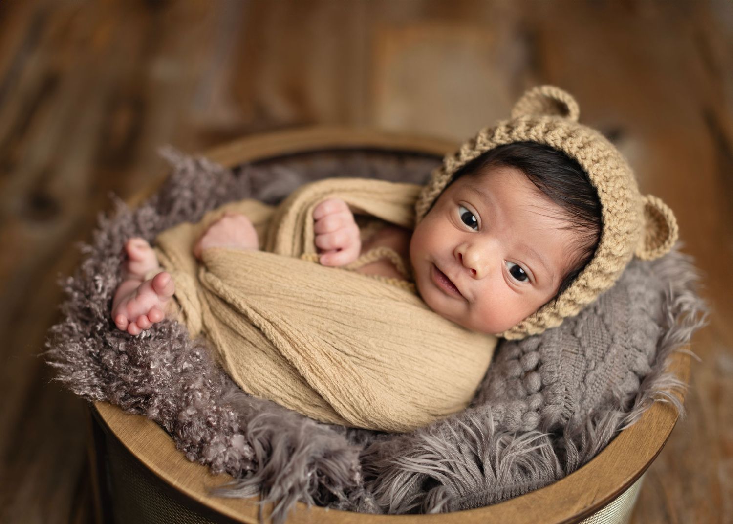 Newborn baby wrapped in yellow swaddle wearing bear bonnet, resting in rustic wooden bucket with gray fur. Elizabeth Lang Photography Surrey