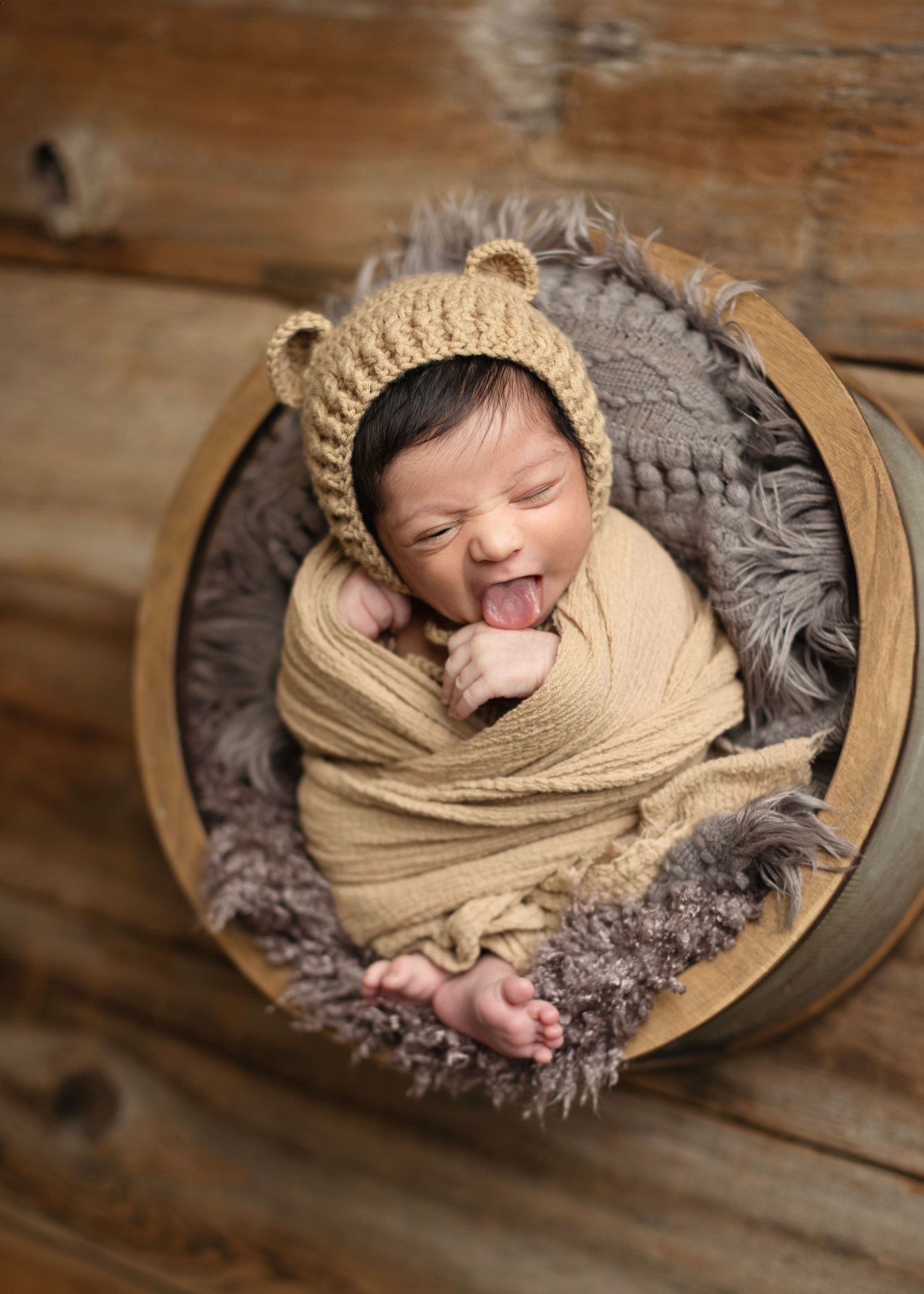Smiling newborn baby wrapped in tan swaddle wearing bear ear bonnet hat, nestled in wooden bucket with gray fur. Elizabeth Lang Photography