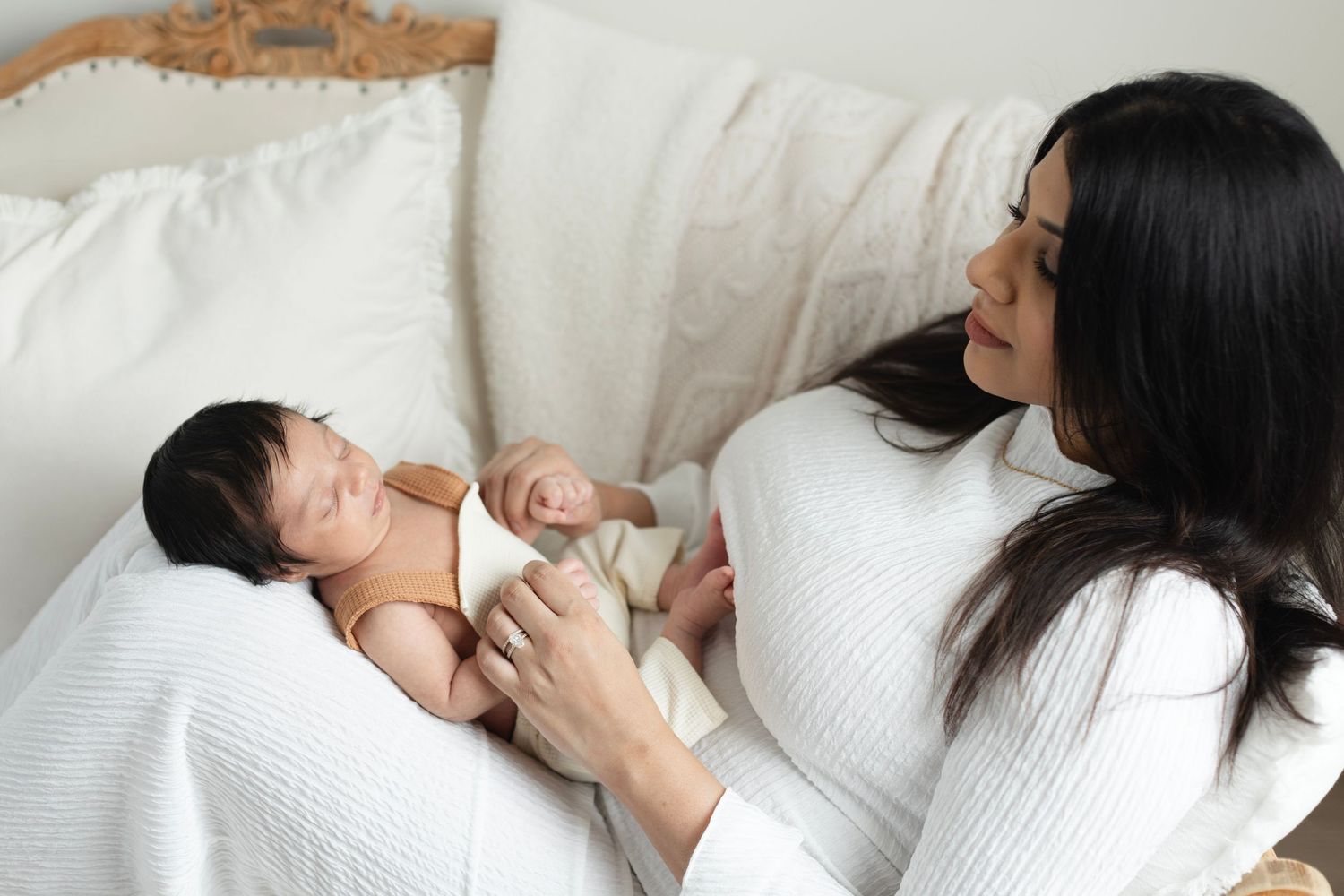 A mother in white gazes lovingly at her newborn baby lying on a white bed. Elizabeth Lang Photography Surrey