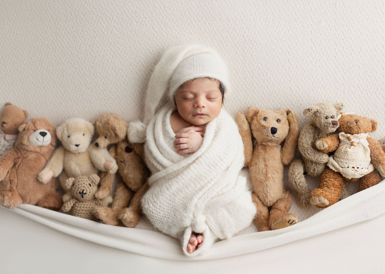 Newborn baby wrapped in white swaddle sleeping peacefully surrounded by soft teddy bears in a professional photo. Elizabeth Lang Photography