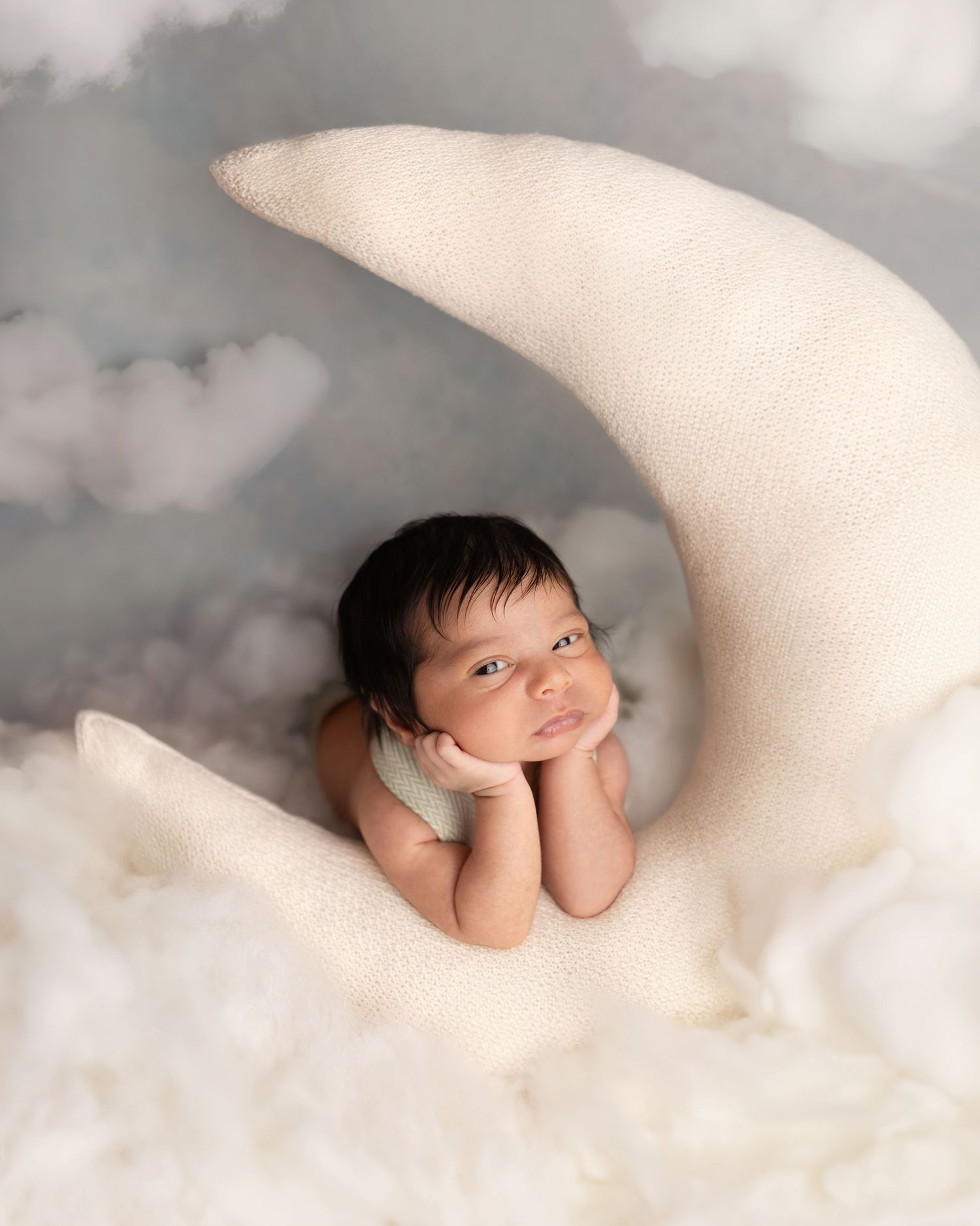 Newborn baby resting chin on hands atop a cream moon prop with soft clouds in a dreamy studio photo. Elizabeth Lang Photography Surrey