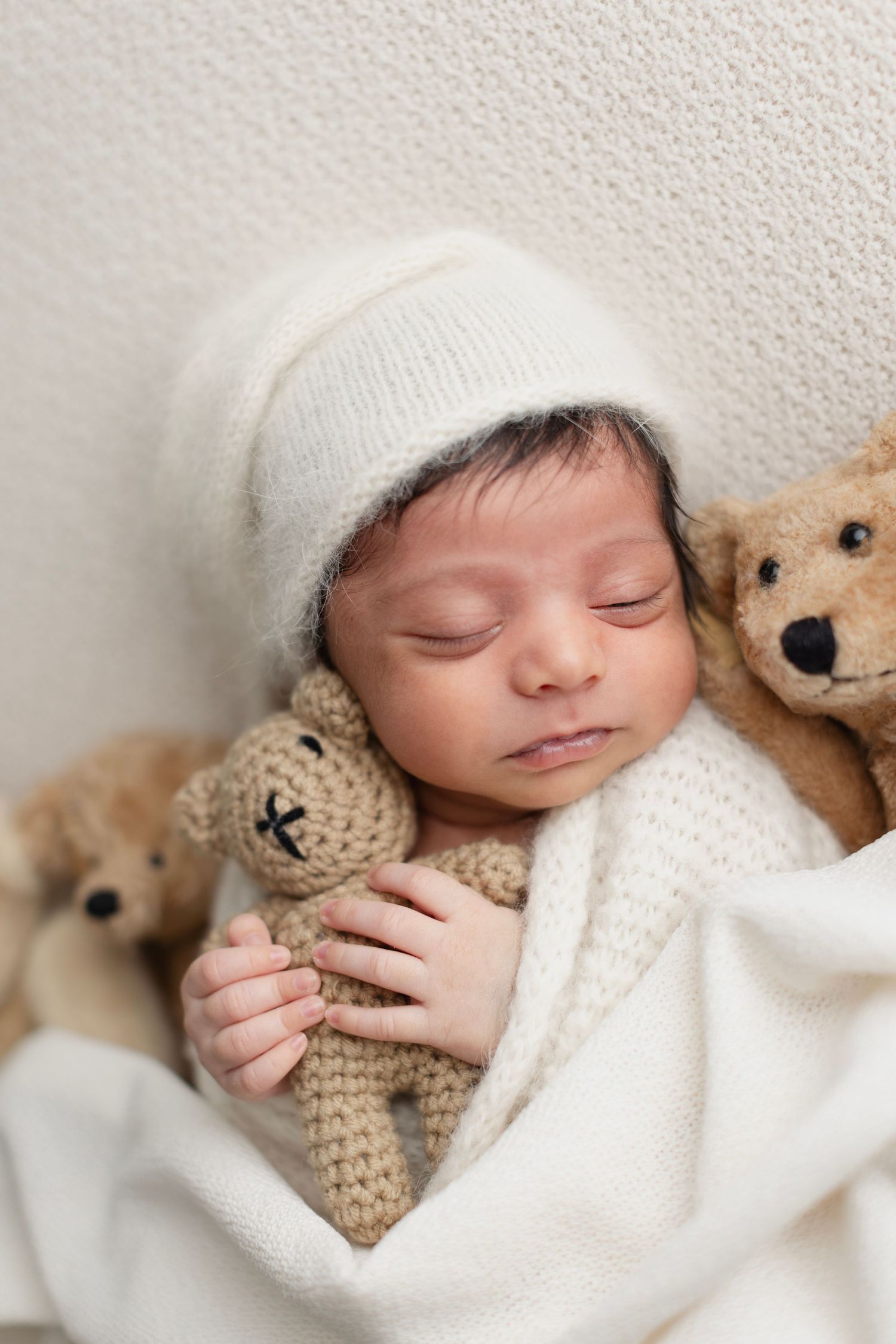 Sleeping newborn baby in white knit hat holding a small teddy bear, wrapped in a soft white blanket. Elizabeth Lang Photography Surrey