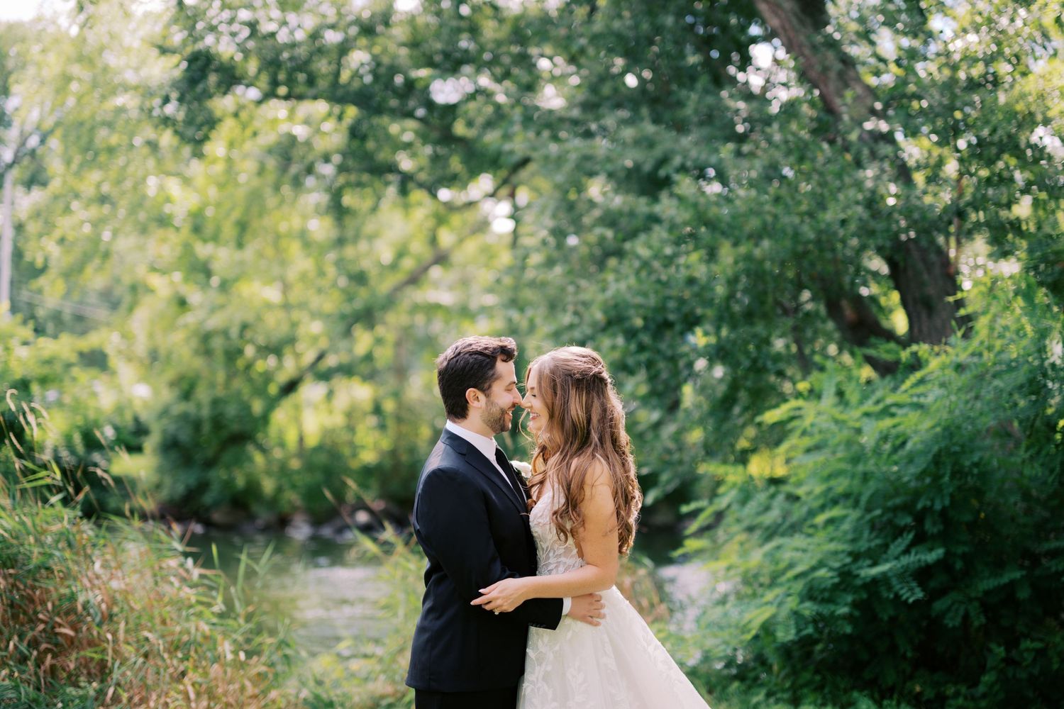 Bride and groom share romantic moment near stream surrounded by lush green trees on wedding day.