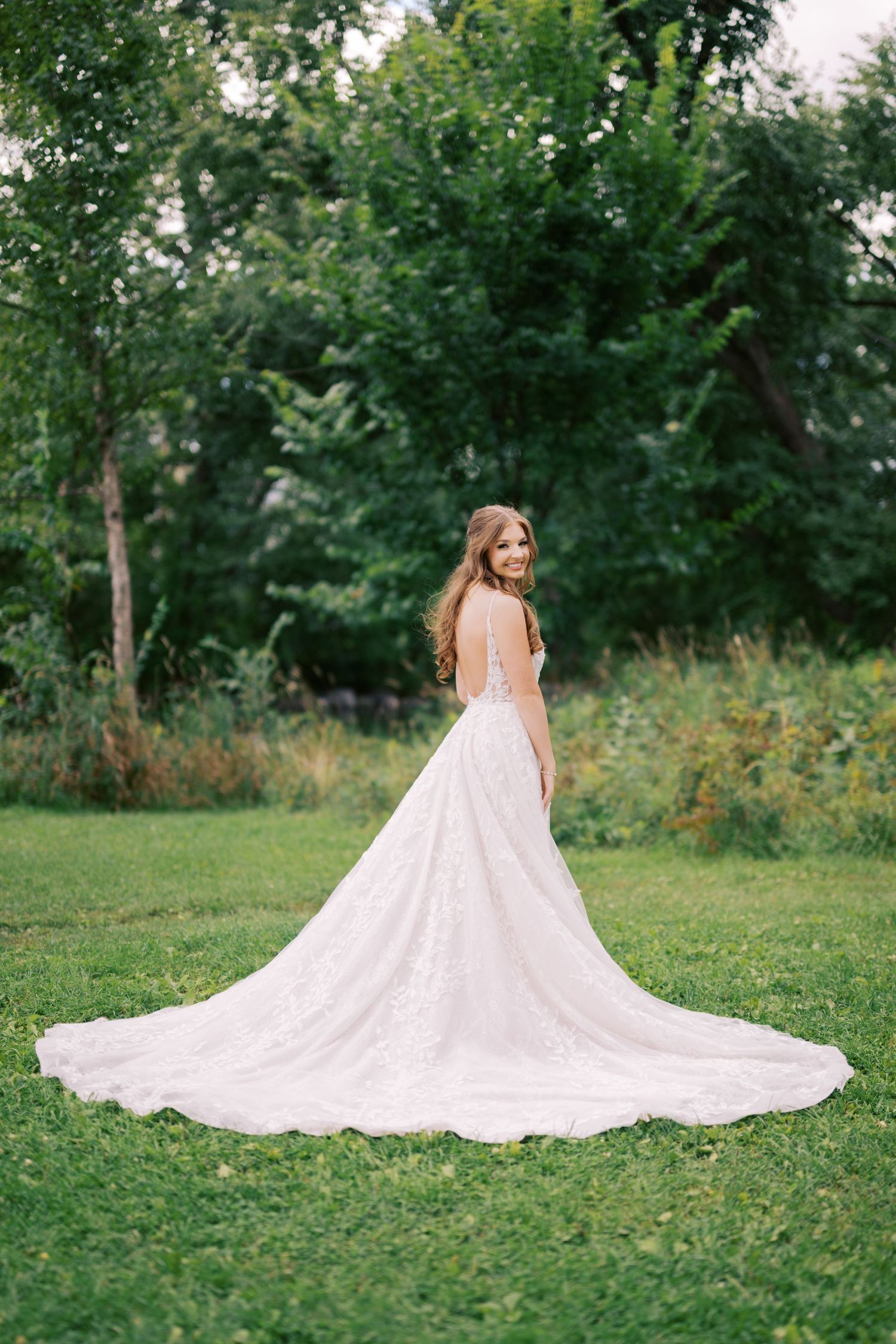 Bride in elegant lace wedding gown with long train standing in green garden turning to smile.