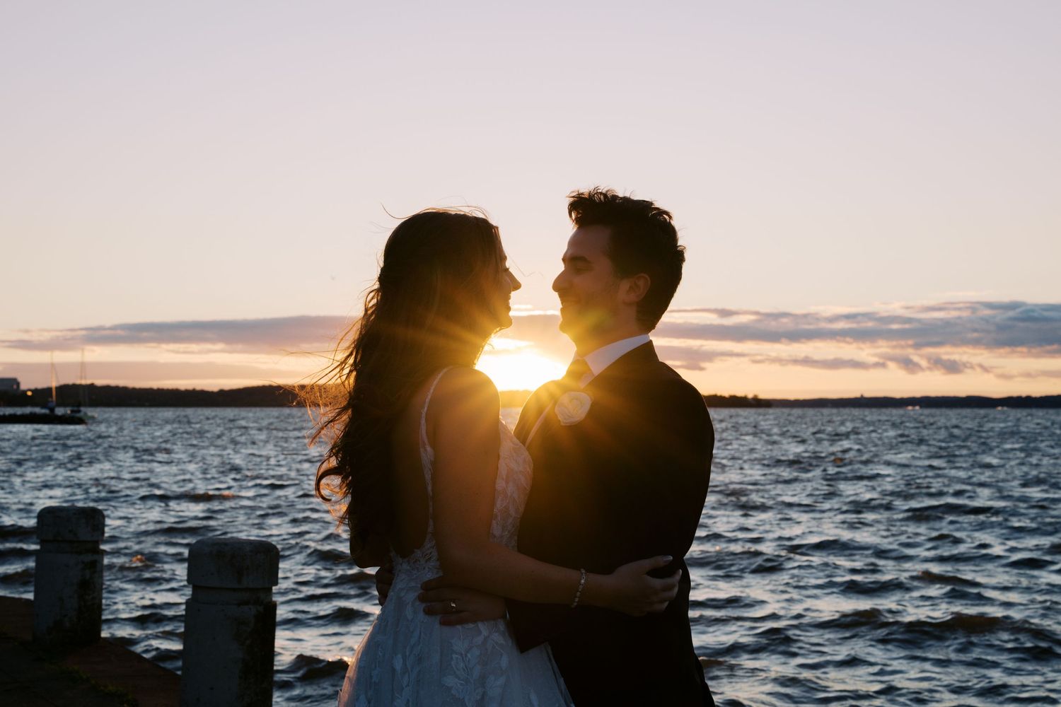 Couple embracing at sunset by the waterfront in wedding attire, silhouetted against golden sky.