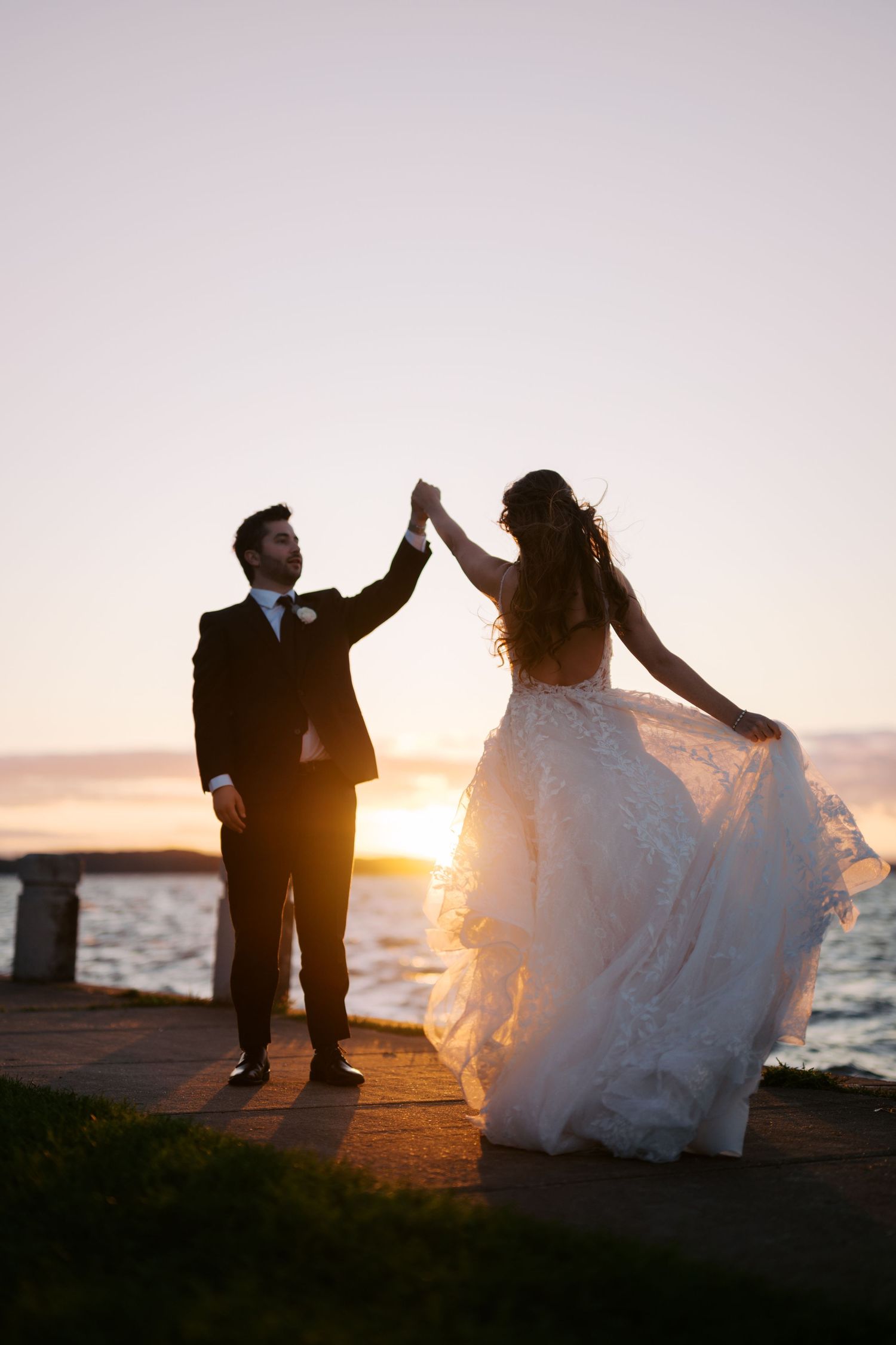 Bride and groom dance at sunset by the water, groom spinning bride in flowing lace wedding gown.
