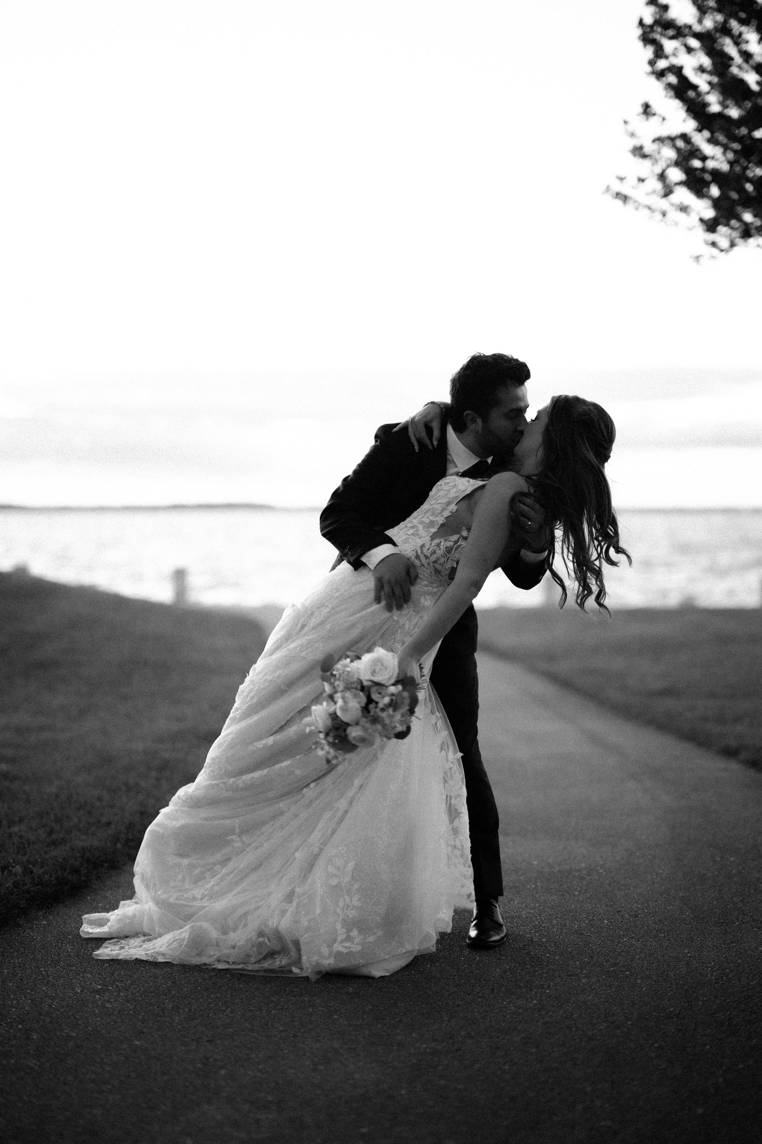 Black and white photo of groom dipping bride for a kiss on pathway beside open water at sunset.