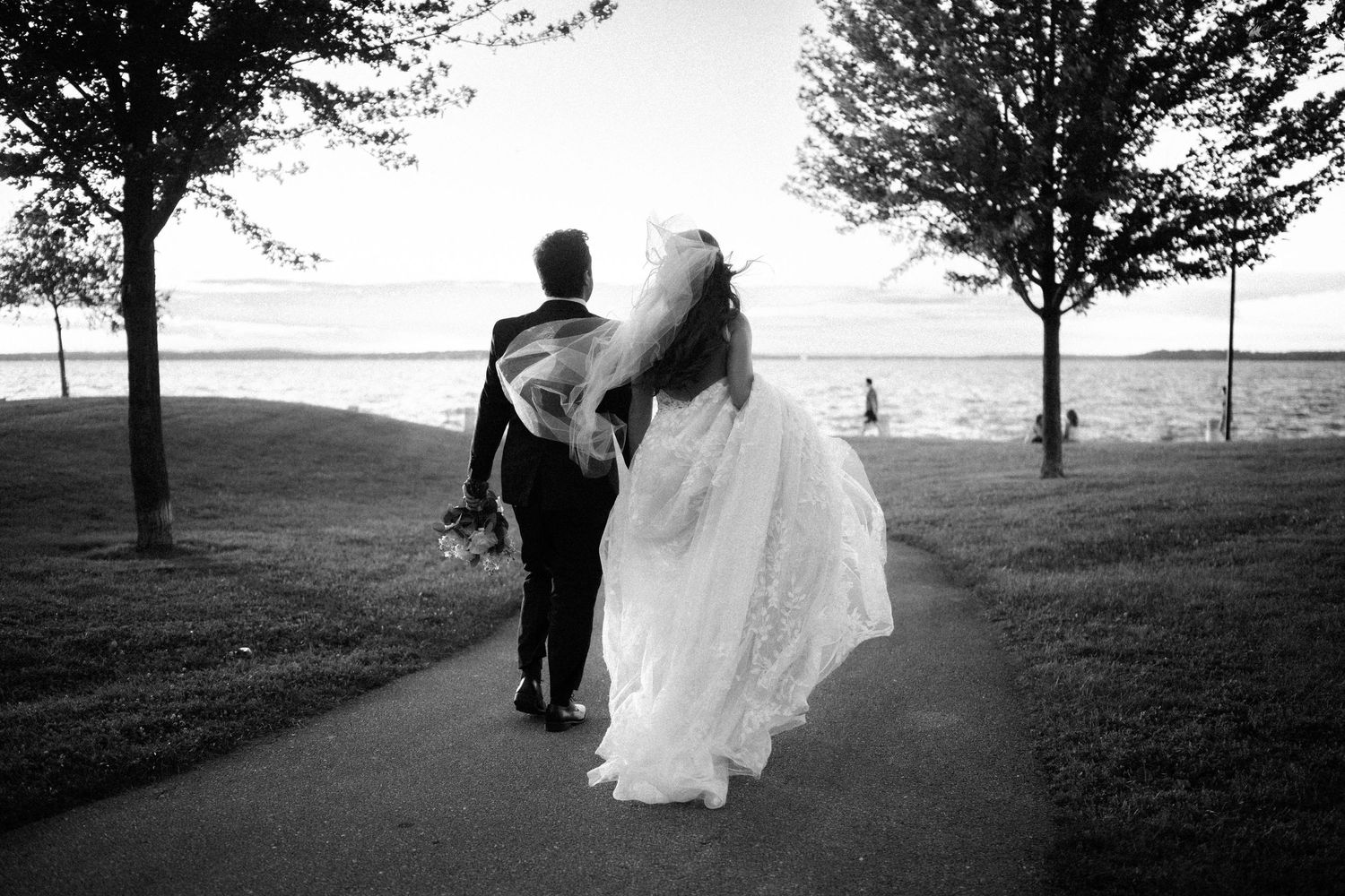 Black and white photo of bride and groom walking hand in hand along a path toward a scenic waterfront.