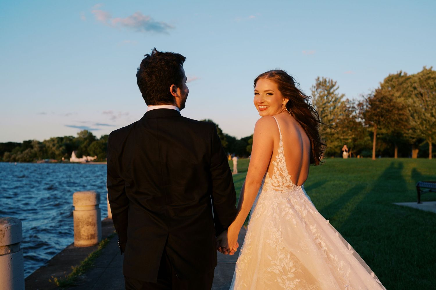Bride and groom hold hands walking by lakeside at golden hour, bride glancing back over shoulder.