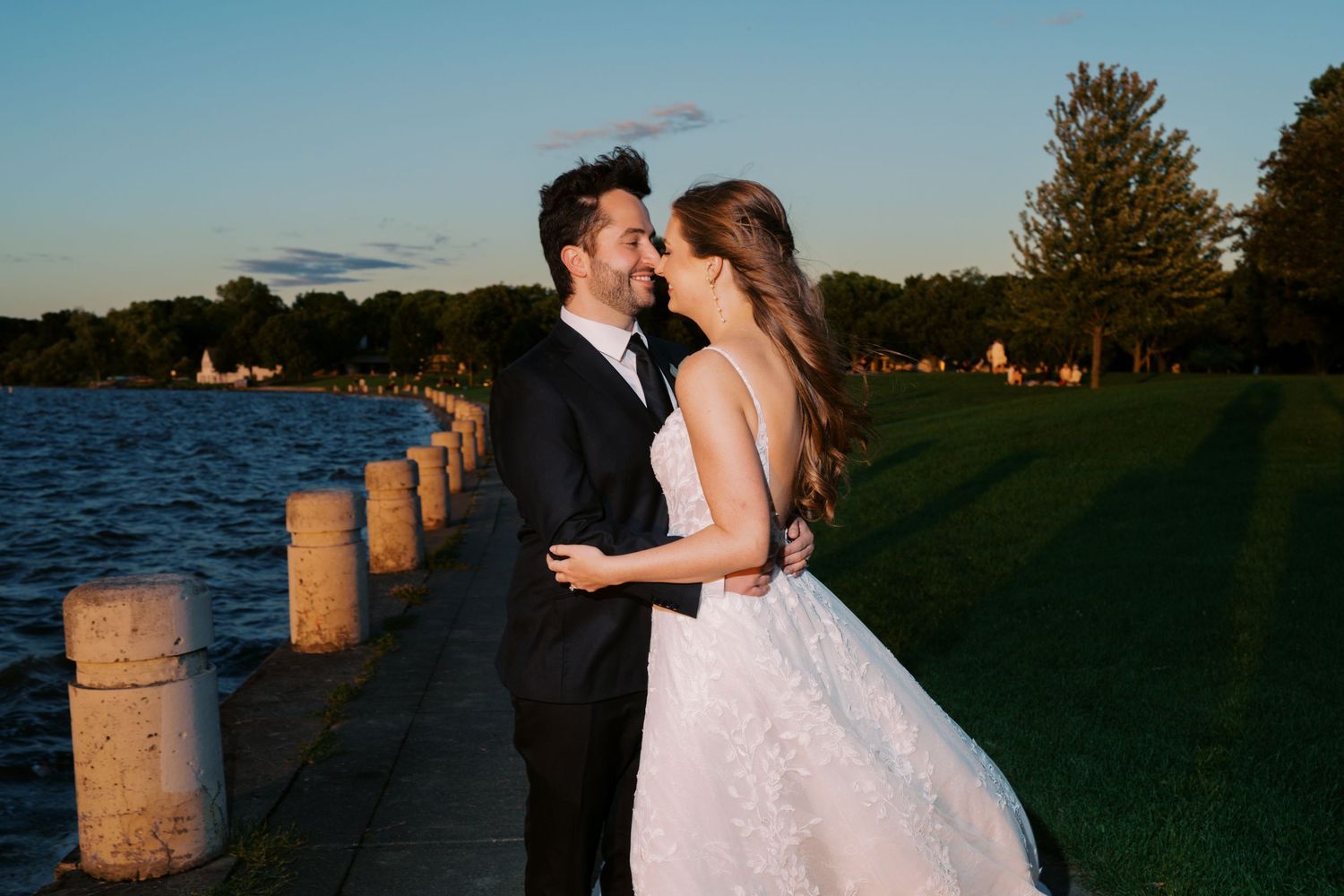 Bride and groom sharing a romantic moment by a sunlit waterfront, embracing during golden hour at their wedding.