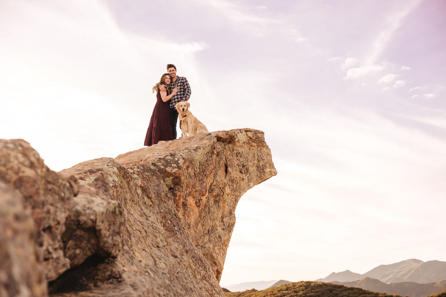 Couple and golden retriever standing on rocky mountain summit with dramatic sky backdrop.